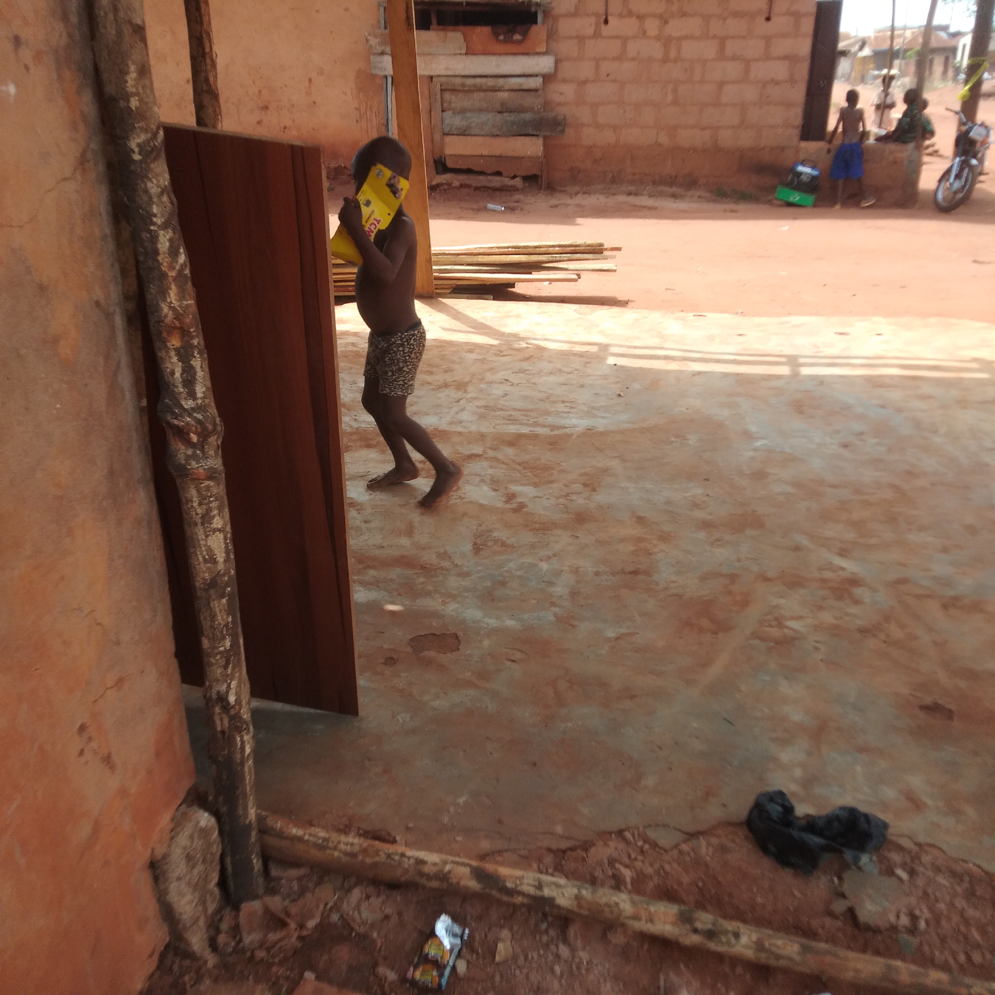 A young, dark-skinned child is walking, barefoot, across a red-earth courtyard. He is holding a yellow rectangular object with lettering on it. In the background, two figures are sitting on a low, brick construction. A motorcycle is parked next to them. The location appears to be a rural or impoverished area of a city. Discarded packaging, including plastic bags and what appears to be food wrapping, litters the ground. The location is in the city of Unknown, Unknown.