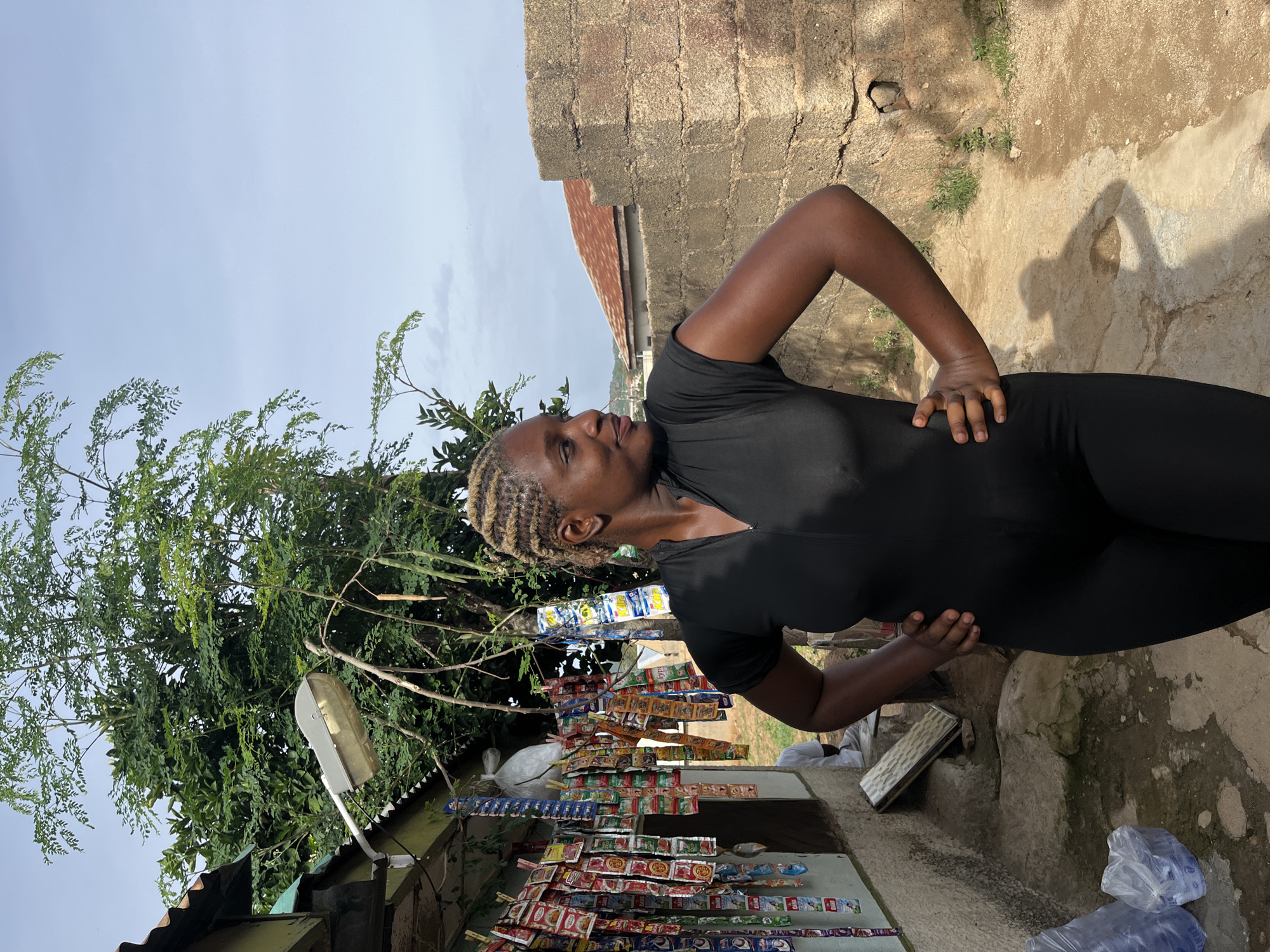 In the city of Rafin Par, Nigeria, a woman with blonde-streaked braided hair stands with her hands on her hips, facing away from a small roadside shop. She is wearing a black, short-sleeved, full-body garment. The shop, constructed from light-colored materials with a green-painted overhang, displays numerous small, colorful packets of products hanging from strings. Behind the shop, a large tree with green leaves and some hanging pods is visible, along with a street lamp. To the right of the woman, a rough, textured brick wall forms the edge of the scene, with a rusted metal roof visible in the background. The ground is uneven, composed of dirt and scattered stones, with a pathway leading away. In the foreground, plastic-wrapped bottles of water are placed on the ground. The sky above is a light blue with some scattered clouds.