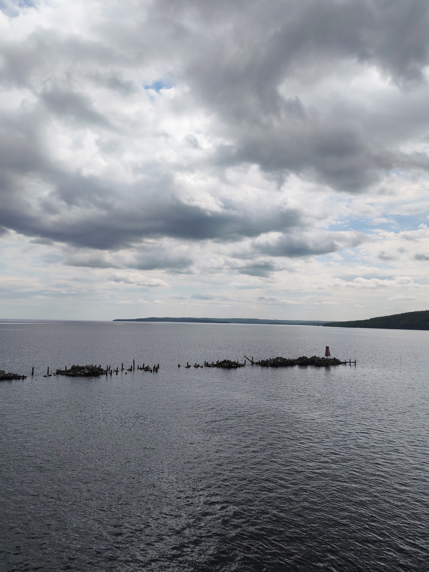 The image presents a broad, tranquil view of a large body of water, likely a lake or bay, under a vast, cloudy sky near Medvezhyegorsk, Russia. The sky dominates the upper portion of the frame, filled with a dense, textured array of grey clouds, interspersed with brighter patches and faint glimpses of blue sky, suggesting an overcast but not actively rainy day.
The water, appearing dark slate grey with subtle ripples, occupies the majority of the scene. Stretching across the mid-ground is a long, low-lying structure composed of rocks and numerous dark, weathered wooden posts protruding vertically from the water. This structure resembles a partially submerged breakwater or the remains of an old pier. On the right side of this rocky formation, a small, distinctive navigational beacon is visible. It is cylindrical with prominent red and white horizontal stripes, designed to guide vessels.
In the far distance, a dark, forested landmass forms a continuous, low horizon line, indicating the opposite shore of the expansive water body. The overall setting conveys a sense of quiet solitude and vastness. The time of day appears to be daytime, under diffuse light. There are no people, explicit activities, or visible text in the image.