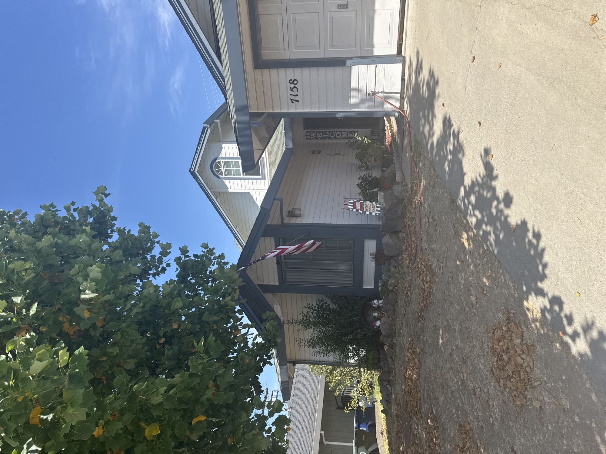 The image shows the exterior of a single-family home located at 7158, Citrus Heights, United States of America. The house is light-colored with a dark-trimmed roof. A two-car garage door is visible on the right side of the house. An American flag is displayed on the front of the house, near a window. The front yard is dry with scattered leaves on the ground. There are several decorative garden gnomes in the front yard. A tree with green leaves shades a portion of the scene. The sky is blue with some wispy clouds.