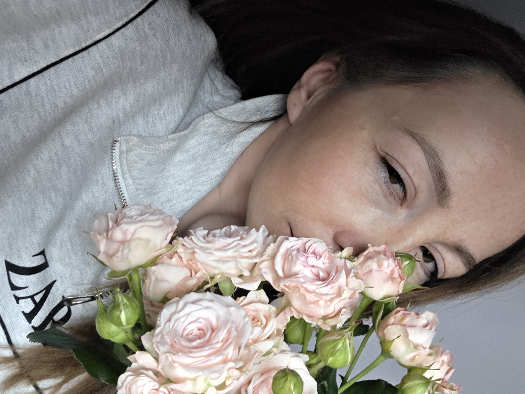 A woman lies on her side, her face close to a bouquet of light pink roses and green buds. She has dark hair and fair skin. She is wearing a light gray zippered sweatshirt with black trim and the word "ZARA" partially visible on the front. The background is a plain gray. There is no indication of this image being taken in the city of Chaikovsky, Russia.