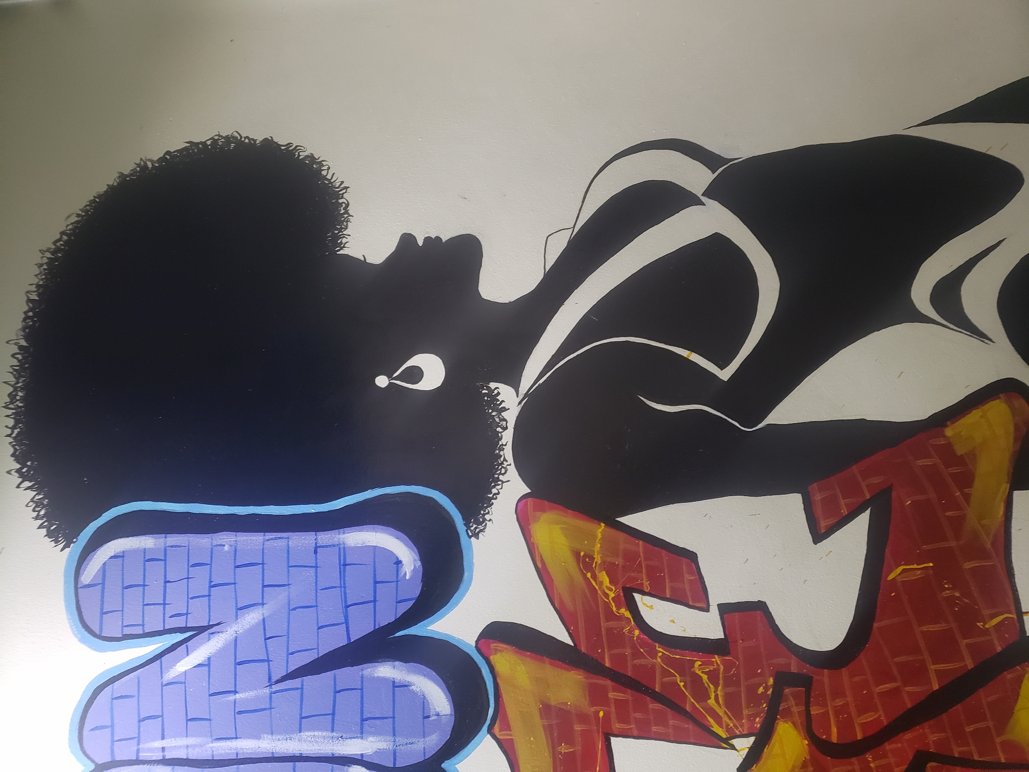 The image is of a section of wall art, possibly graffiti, in what appears to be an indoor location in the city of Adankolo, Nigeria. The focal point is a silhouette of a person with a large Afro hairstyle. The person is adorned with what appears to be an earring. Stylized lines suggest the contours of a body. Below the silhouette are two large graffiti-style letters, the left one colored in shades of blue and the right in shades of red and yellow. The background is off-white.