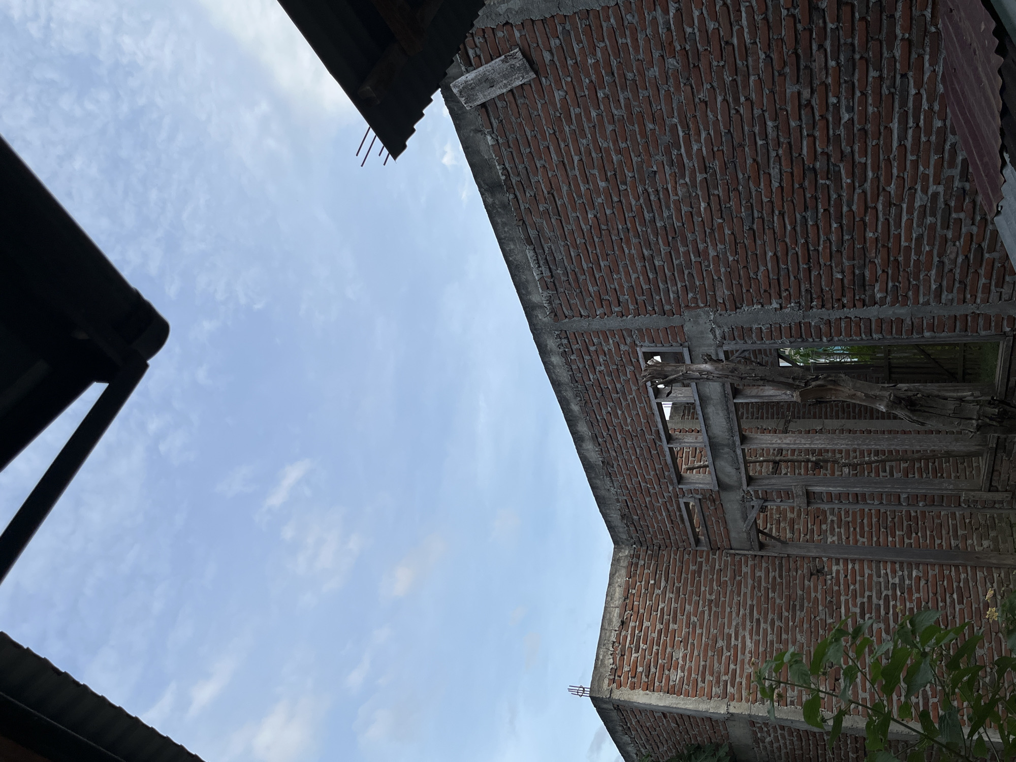 The image shows a low-angle view of a partly constructed or dilapidated building in Lhokseumawe, Indonesia. The structure features exposed red brick walls and concrete support beams. A section of the building is open to the sky, revealing a light blue sky with sparse clouds. A corrugated metal roof is partially visible on the left side of the frame. Some green foliage is visible in the lower right corner.