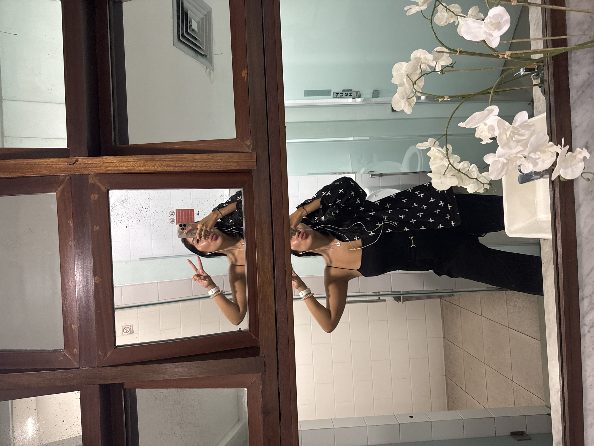 A woman with long dark hair takes a mirror selfie in a brightly lit, tiled restroom. She is wearing a black spaghetti-strap top, black pants with a gold belt, and a black jacket with white cross patterns. She has white earbuds in and is holding a smartphone in her right hand, angled to capture her reflection. Her left arm is raised, and she is making a peace sign. A second, slightly lower mirror reflects a similar view, showing more of the restroom's tiled walls and floor, a toilet, and a frosted glass door. In the foreground of the lower mirror, a vase of white orchids is visible on the counter. The restroom appears to be located in Kuta, Indonesia, suggested by the tropical flora and architectural style.