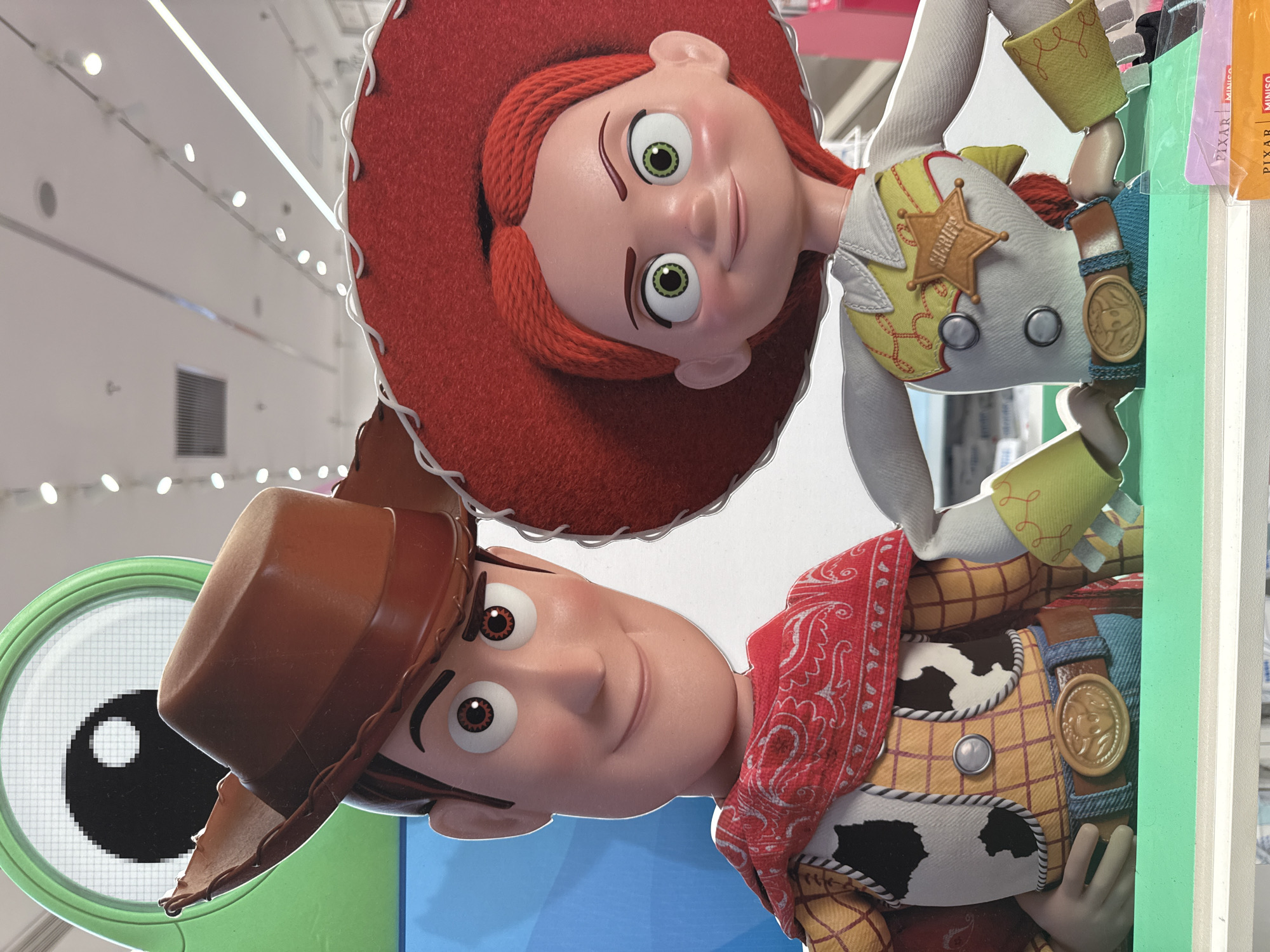 This media file features two Toy Story characters, Woody and Jessie, depicted as life-sized cardboard cutouts. Woody, on the left, is wearing his signature cowboy hat, red bandana, and cow-print vest. Jessie, on the right, is wearing her red cowgirl hat, sheriff's badge, and a yellow and white patterned shirt. Both characters are smiling, with bright, expressive eyes. Woody's eye on the left has a magnifying glass overlay. The background appears to be an indoor setting with white walls and bright ceiling lights, possibly a store or an exhibition space. Some packaging with text is visible on the right edge of the image, including "PIXAR MINISO". The overall mood is cheerful and playful, characteristic of the Toy Story franchise. The image is oriented vertically, with the characters appearing upside down. The scene is likely set during daytime, given the bright lighting. The location context is Xiaoshan District, China.