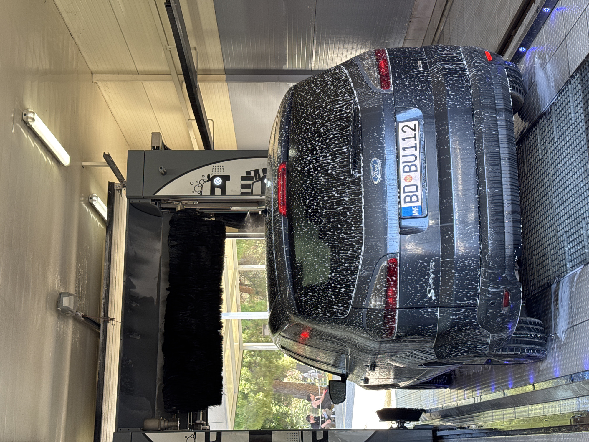 The image depicts a dark-colored Ford S-Max being washed in an automated car wash. The car is covered in white soap foam. The license plate reads "BD BU112," indicating the vehicle is registered in Budva, Montenegro. A large, rotating black brush is visible on the left side of the frame. Outside the car wash structure, green trees and foliage are partially visible. Two people can be seen standing outside. The car wash interior is illuminated by fluorescent lights.