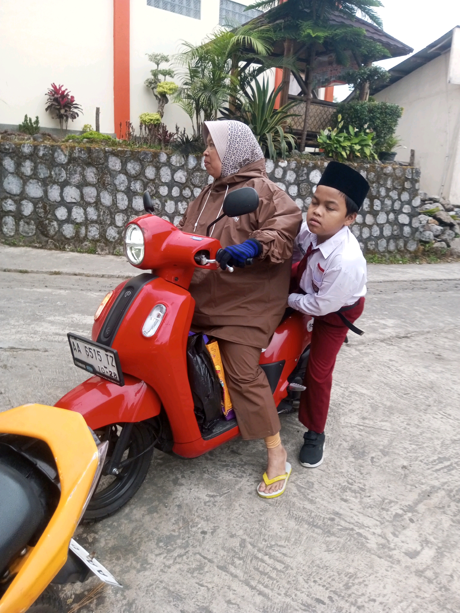 In Kalibeber, Indonesia, a woman wearing a brown hooded jacket and a patterned headscarf is seated on a red scooter. A young boy, dressed in a white school shirt with a red tie and maroon pants, is perched behind her. The scooter has a license plate reading "AA 6515 TZ". The background features a stone wall topped with various plants and a thatched-roof gazebo. The scene is set on a paved surface, likely a road or pathway.