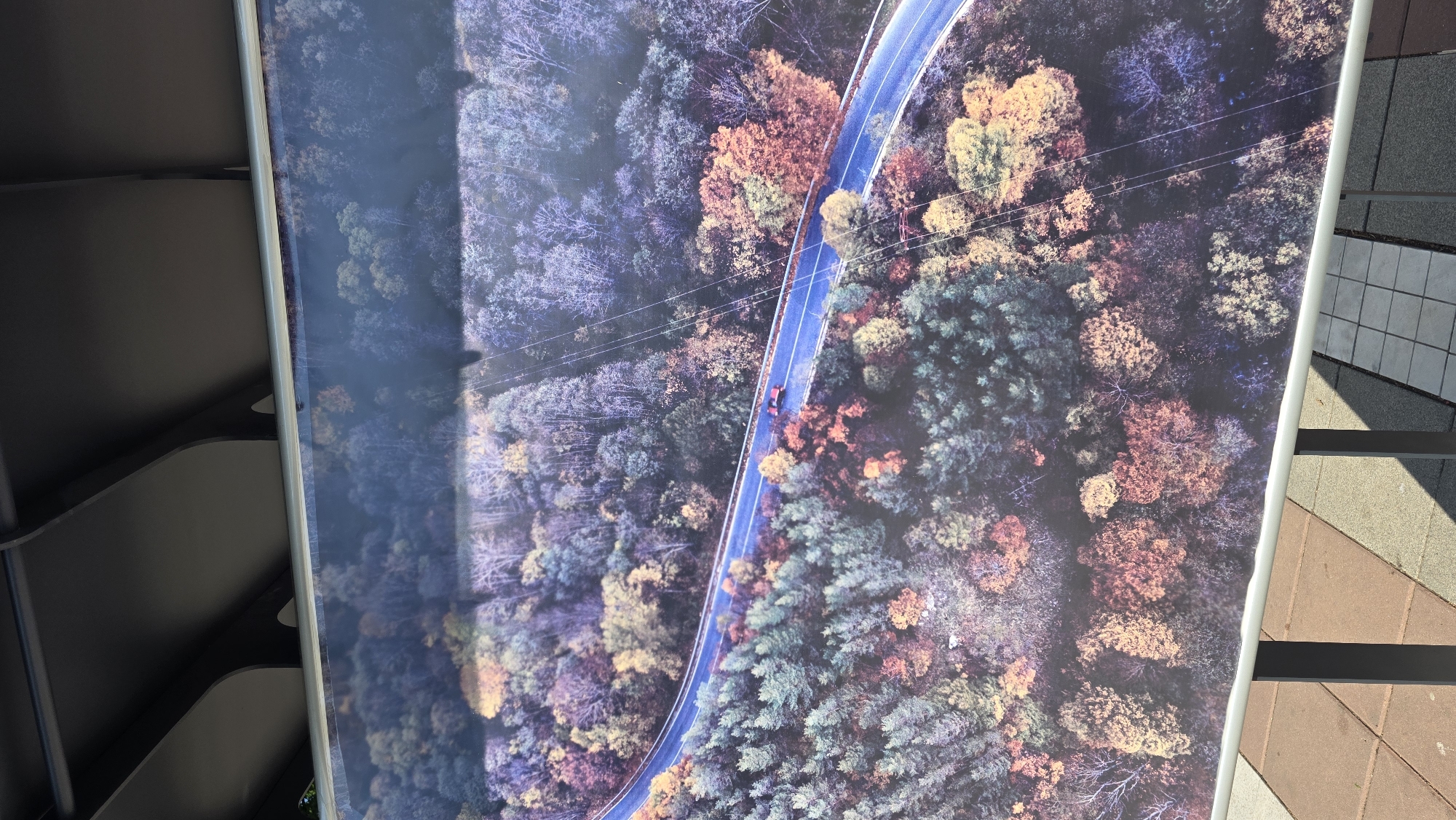 The image is an overhead view of a winding road cutting through a dense forest with trees exhibiting autumn colors. The road is mostly straight but curves slightly. A red car is seen driving on the road. The surrounding forest is a mix of green, yellow, orange, red, and purple trees, suggesting it is autumn. There are also some utility poles with lines visible. The picture is likely part of a larger display. It is unknown if the location is in Blagoevgrad, Bulgaria.