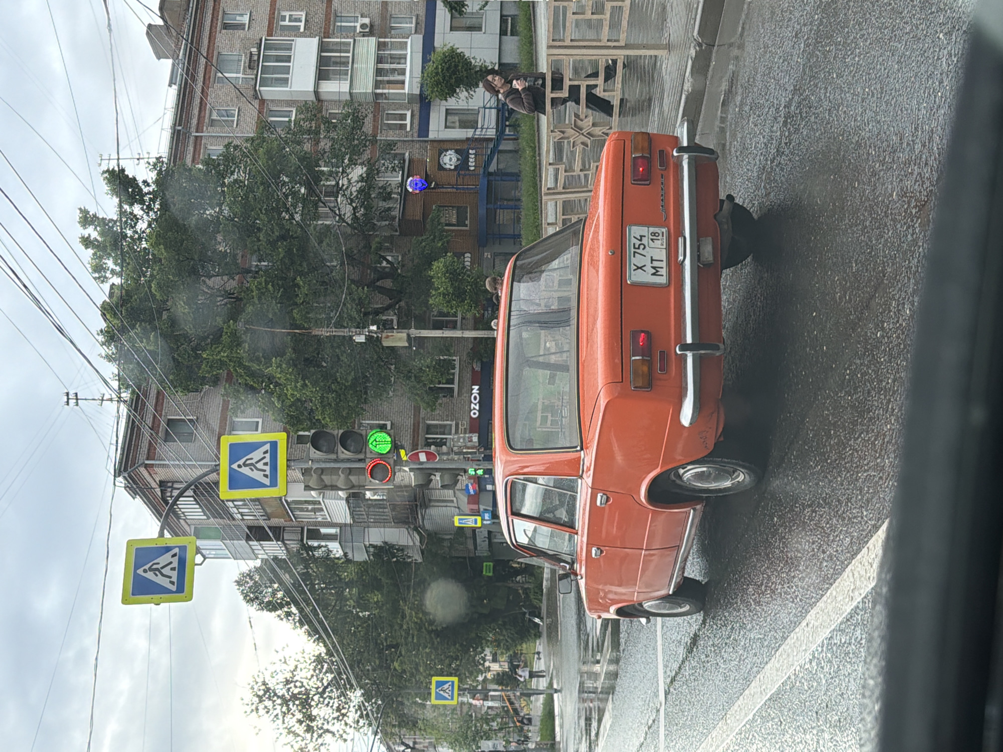 The image shows a street scene in Izhevsk, Russia, on a wet day. A rusty, orange VAZ-2106 car is parked on the right side of the road. A pedestrian crosswalk sign is visible, and traffic lights show a green light. Buildings are visible in the background, including one with an "OZON" sign. A woman is seen standing near a decorative fence. The license plate of the VAZ-2106 car is X 754 MT 18.