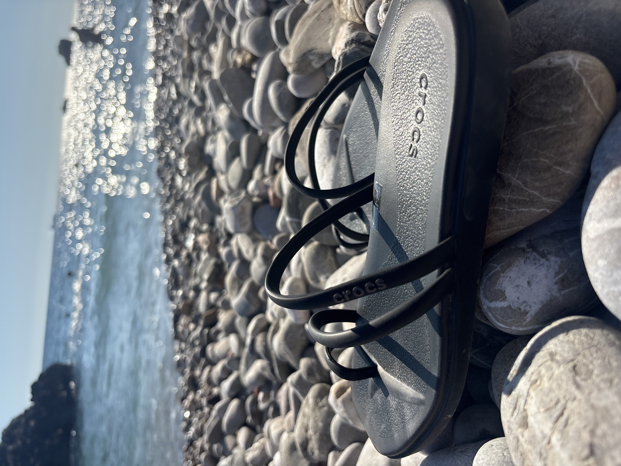 A pair of black Crocs sandals rests on a pebble beach in Bar, Montenegro. The sandals are placed on a bed of smooth, rounded grey and white pebbles of varying sizes. In the background, the Adriatic Sea sparkles with reflected sunlight, slightly out of focus. A portion of the blue sky is visible in the upper left corner.