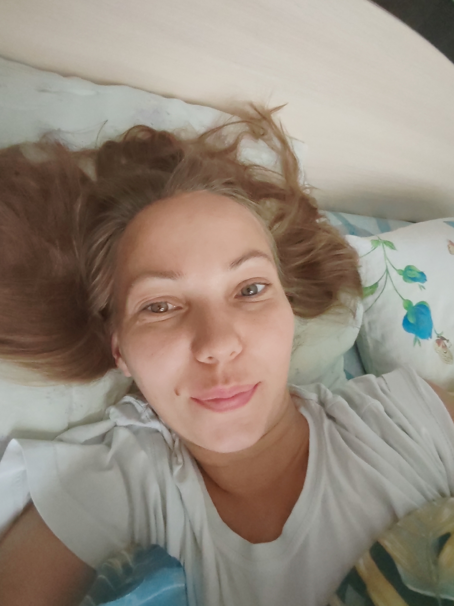 A selfie-style, overhead shot captures a woman lying in bed, her head turned towards the camera. She has fair skin, light brown hair spread out around her head, and light brown eyes. Her lips are slightly parted, and she has a small mole on her left cheek. She is wearing a white t-shirt. The background consists of a pillow with a floral pattern featuring blue flowers and green leaves, and a light-colored headboard. The setting appears to be a bedroom. The image does not provide any specific location information for Чухлэм, Russia.