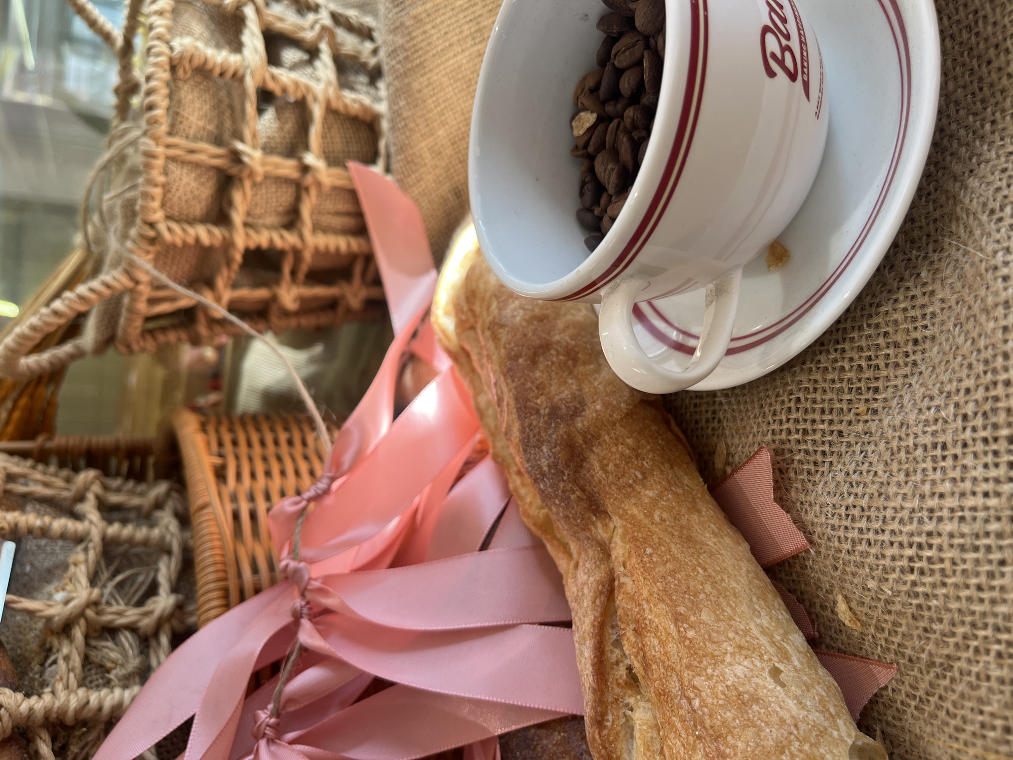 The image shows a gift basket containing coffee beans in a white and red rimmed coffee cup atop a saucer, a baguette, and pink ribbons, possibly in Bắc Nha Trang, Vietnam. The basket is made of natural materials, with a mix of woven and netted sections. The background consists of a burlap-like fabric.