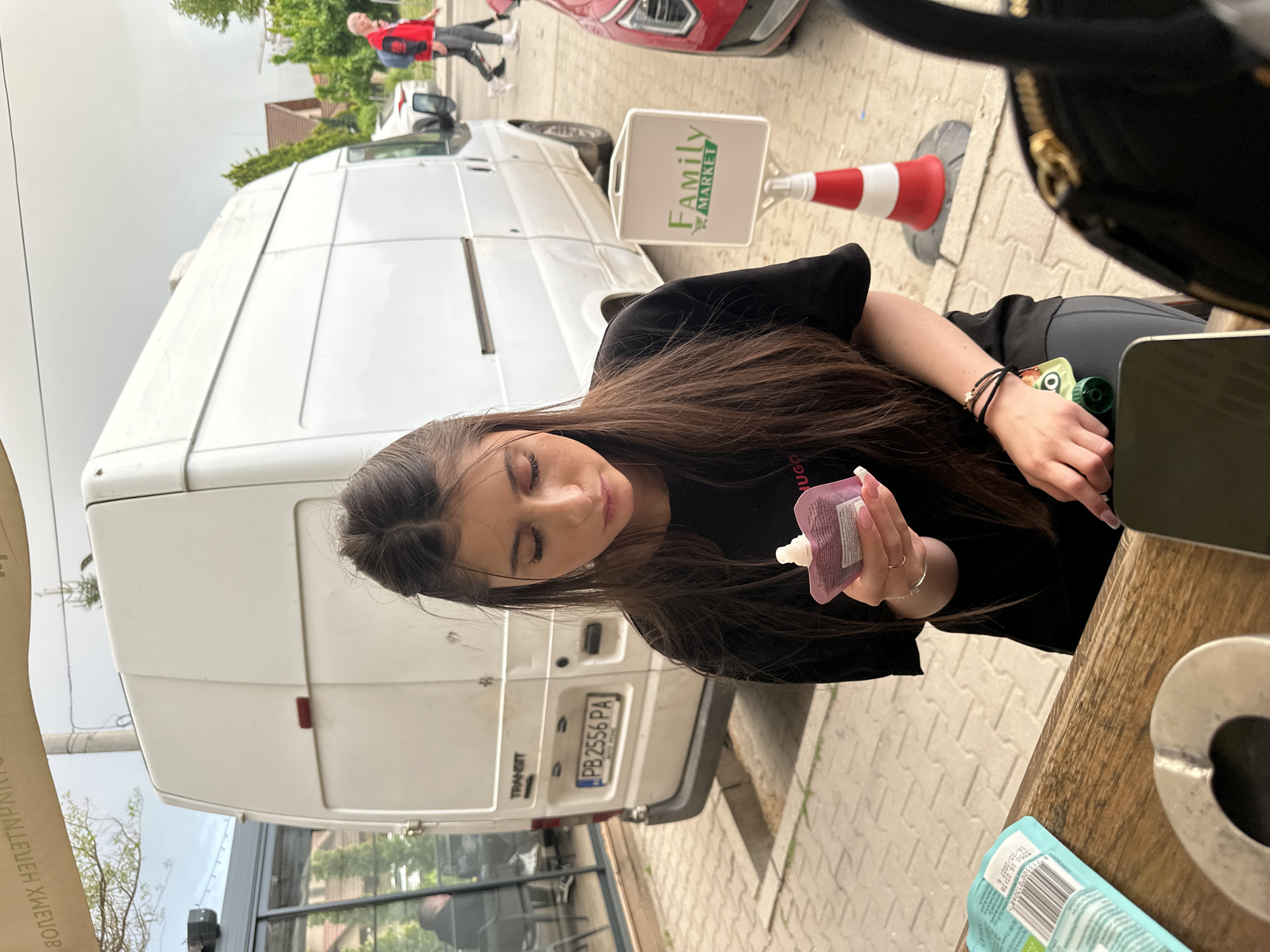A medium-shot image taken at street level depicts a young woman sitting at a table outdoors. She is holding a lavender-colored tube in her hands. She is wearing a black t-shirt with the word "HUGO" printed on it and has long dark hair. She is also wearing black bracelets on her left wrist. The table she sits at is made of light-colored wood. On the table, next to her, is a cell phone and an ashtray. In the lower left corner of the image, there is a cardboard food container.
In the background, there is a white van parked. The van's license plate reads "PB 2556 PA". Next to the van, there is a sign that reads "Family MARKET".
In the far background, pedestrians walk by. The photograph was taken in Plovdiv, Bulgaria.