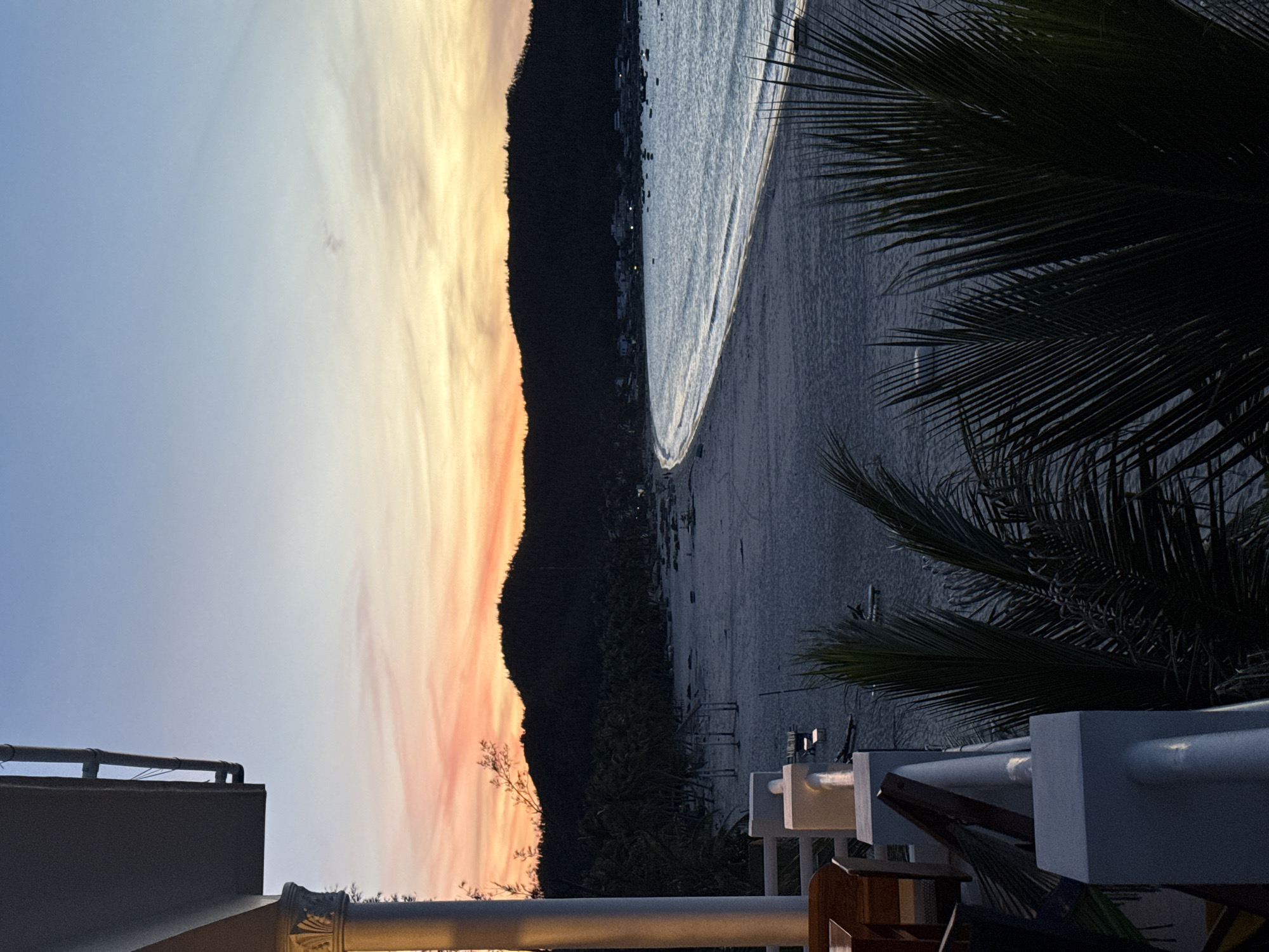 The image captures a serene coastal scene at dusk. The sun is setting, casting a warm, orange and pink glow across a vast sky filled with wispy clouds. The silhouette of a dark, tree-covered hill stretches horizontally across the frame, separating the sky from the beach and ocean. The ocean itself is a deep, dark blue, with gentle waves lapping at the sandy shore. In the distance, a small, dimly lit town or village is visible along the coastline, with scattered lights twinkling in the fading light.
In the foreground, the dark fronds of a palm tree frame the right side of the image, adding a tropical feel. Below the palm tree, a white railing and a portion of a building are visible, suggesting a balcony or terrace overlooking the beach. There are also glimpses of furniture, including chairs and a table, further indicating a resort or residential setting. The overall mood is peaceful and tranquil, evoking a sense of calm as day transitions into night. The time of day is clearly sunset or twilight. There is no visible text in the image.