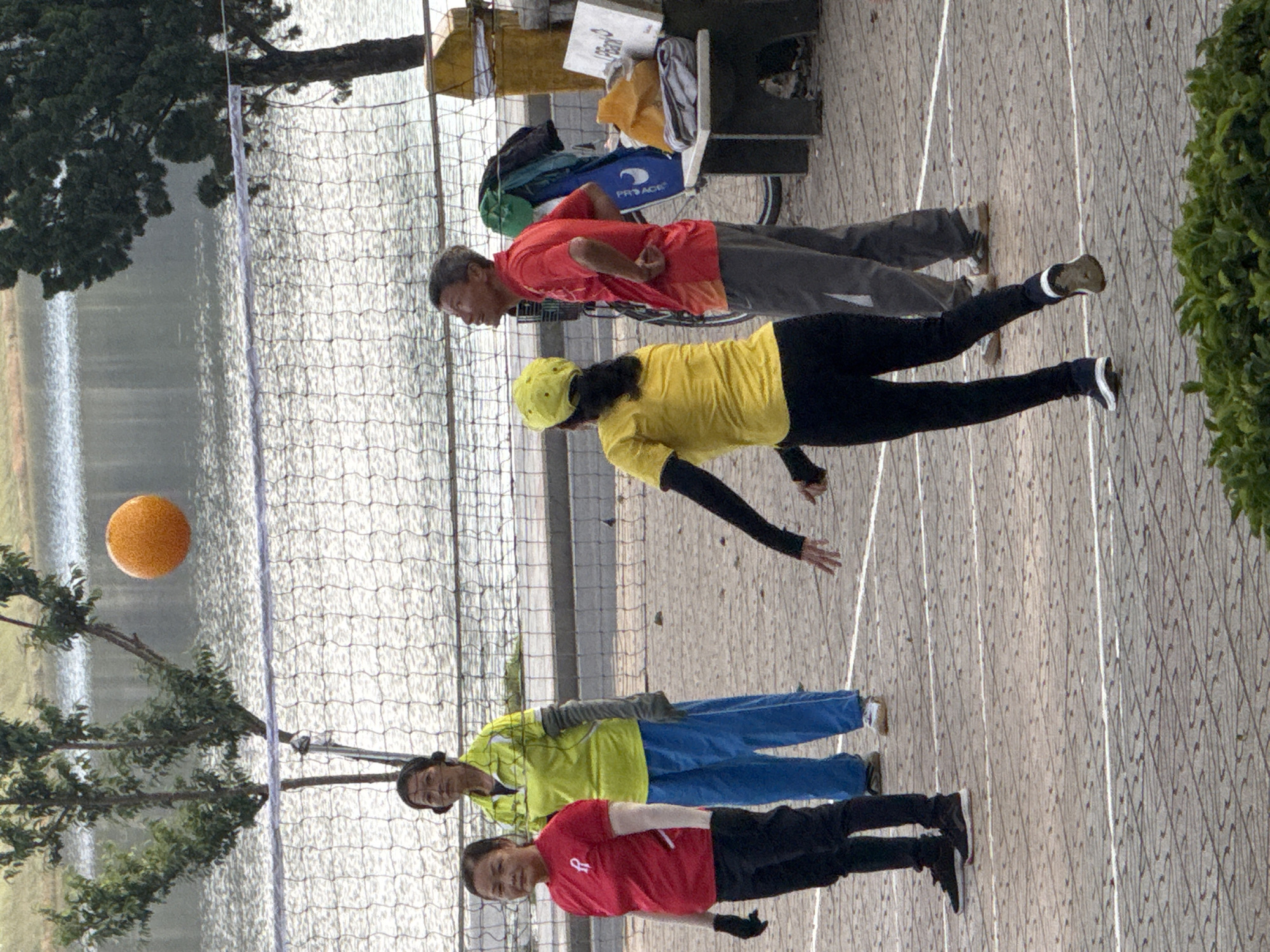 The image depicts a group of four individuals playing volleyball on a paved court adjacent to a body of water. The setting appears to be Da Lat, Vietnam, given the typical mountainous landscape and the style of clothing. In the foreground, two women and two men are actively engaged in the game. The woman on the left wears a red t-shirt and black pants, while the woman next to her is in a lime green shirt and blue pants. The two men are on the right side of the court. One man is wearing a red t-shirt and grey pants, and the other is in a yellow t-shirt and black pants, with a yellow cap. A bright orange volleyball is suspended in the air above the net, suggesting the game is in progress. The background features a net stretching across the court, a calm body of water, and trees on the far bank. The ground is paved with interlocking bricks, and white lines mark the boundaries of the court. A bench and a trash bin are visible to the right of the court, near a tree. The overall atmosphere is one of a casual outdoor sporting activity.
