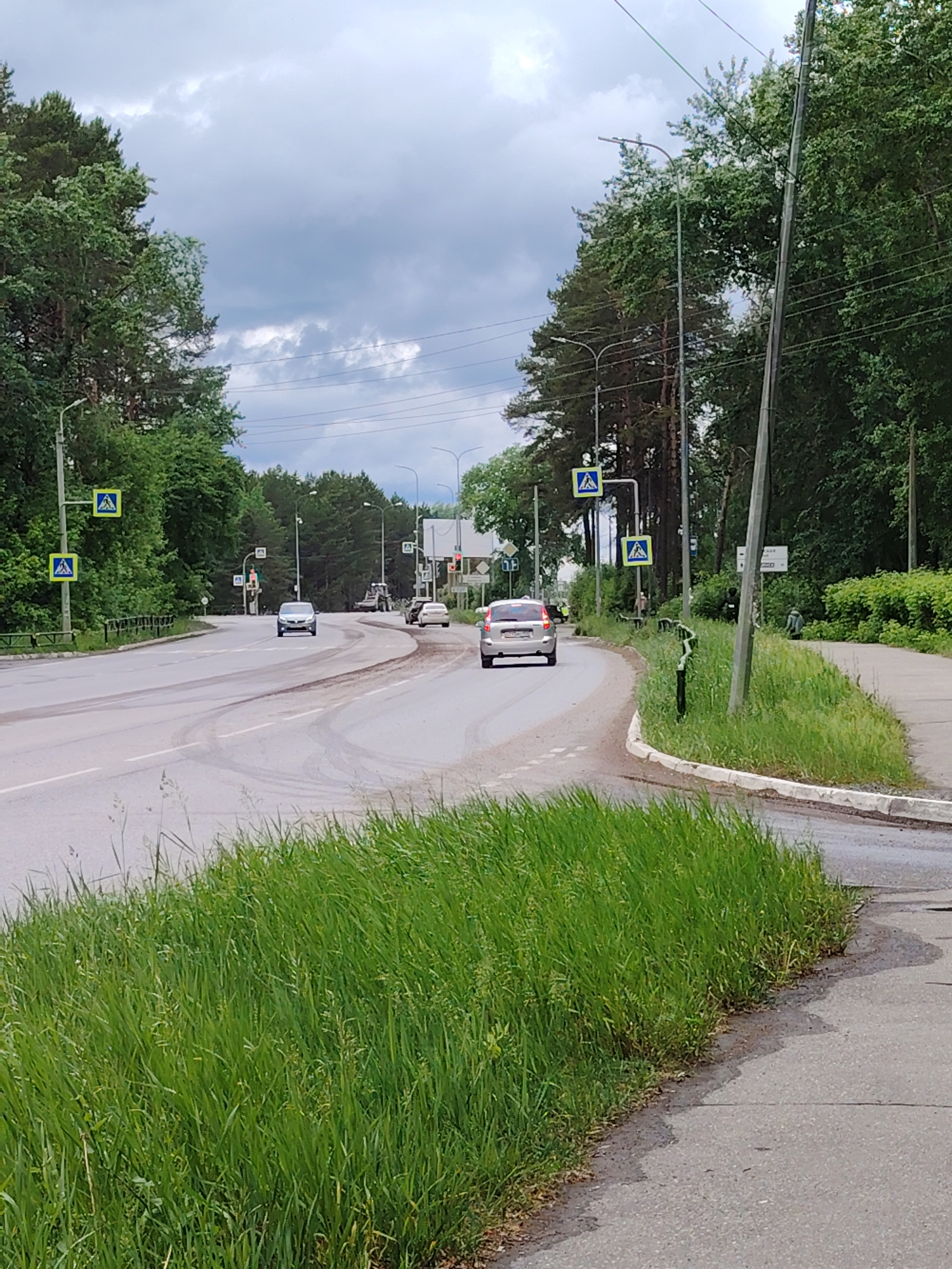 The image shows a street scene in the city of Chaikovsky, Russia. The view is taken from the side of a road, looking towards a slight bend with three cars visible in the distance. The road appears damp with tire tracks. Green trees flank both sides of the road. Roadside pedestrian crossing signs are visible on either side of the road. In the right foreground, there is a patch of green grass with a sidewalk adjacent to the road. Power lines and street lights run overhead. The sky is overcast with dark clouds. There are several people standing on the sidewalk, and the scene suggests an urban or suburban environment.