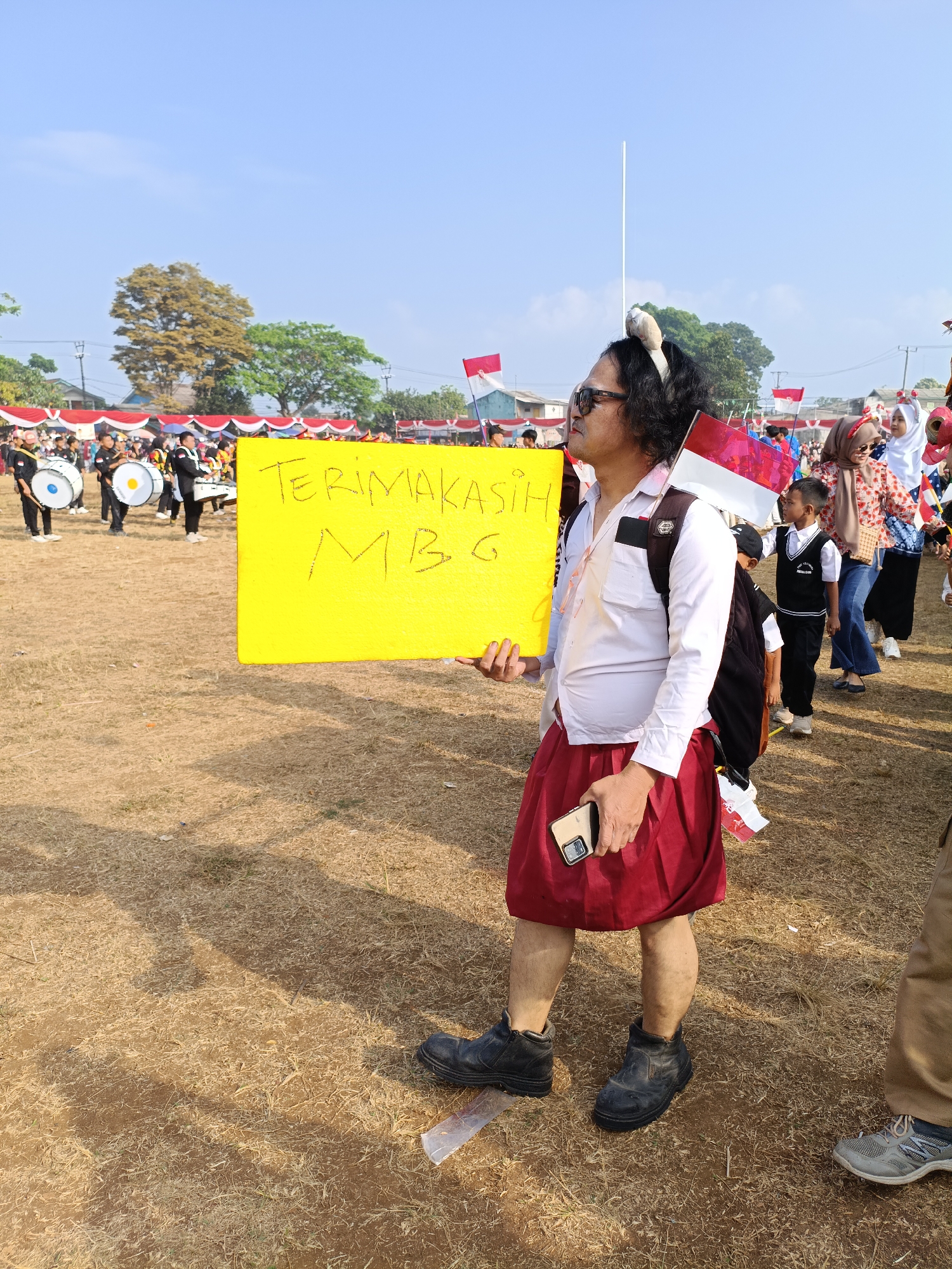 In the city of Cibodas, Indonesia, on a dry, grassy field under a clear blue sky, a person wearing a white shirt, a red skirt, and black boots stands holding a yellow sign that reads "TERIMAKASIH MBG." The individual also wears sunglasses and has a pair of fuzzy animal ears on their head, along with an Indonesian flag tucked into their collar. In the background, a marching band with drums is visible, along with a crowd of people, some of whom are holding small Indonesian flags. The scene appears to be outdoors during a daytime event, possibly a parade or festival.