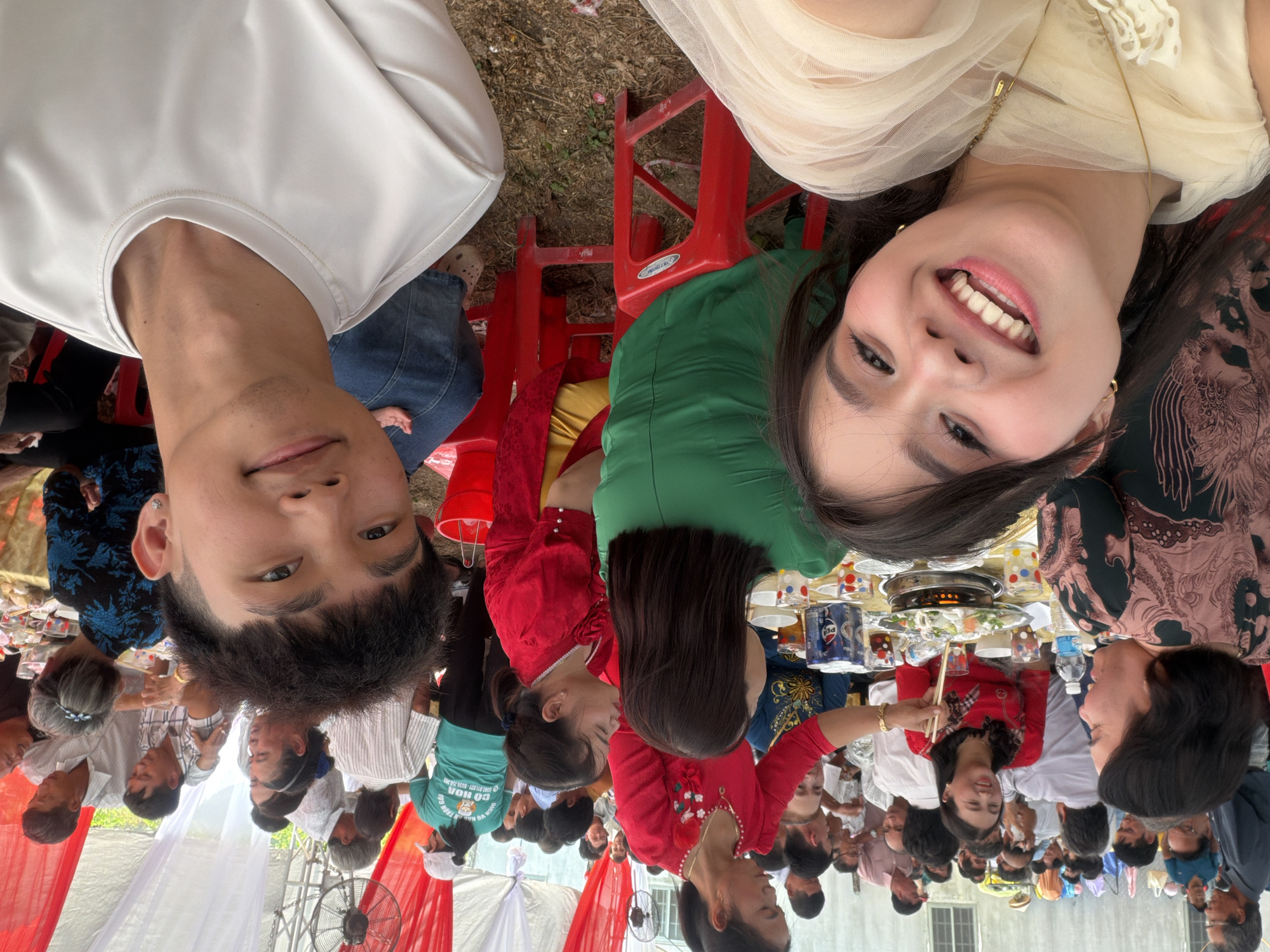 This image depicts a vibrant outdoor gathering, likely a communal meal or celebration, in Tay Son Commune, Vietnam. In the foreground, a man and a woman are taking a selfie. The woman on the right is smiling broadly with an open-mouthed expression, revealing her teeth, while the man on the left has a more subtle smile. Both appear to be in good spirits.
Behind the two main subjects, numerous people are seated at multiple tables arranged under temporary white and red fabric canopies. The tables are laden with food, including what appears to be a hot pot in the center of one table, surrounded by various dishes, cups, and bottles of water. Pepsi cans are also visible on the tables. Attendees are engaged in eating, conversing, and socializing. The chairs are predominantly red plastic, with at least one bearing the visible brand name "DUY TAN".
The event is held outdoors on a patch of ground that looks like dirt or sparse grass. A large fan is visible towards the bottom of the image, suggesting a warm climate. The natural lighting indicates that the event is taking place during the daytime. Visible text on a green t-shirt in the background includes "CỔ HOÁ", "CO HOA", "CHUYÊN VIỆT", and a partial phone number. The overall scene conveys a lively and communal atmosphere.