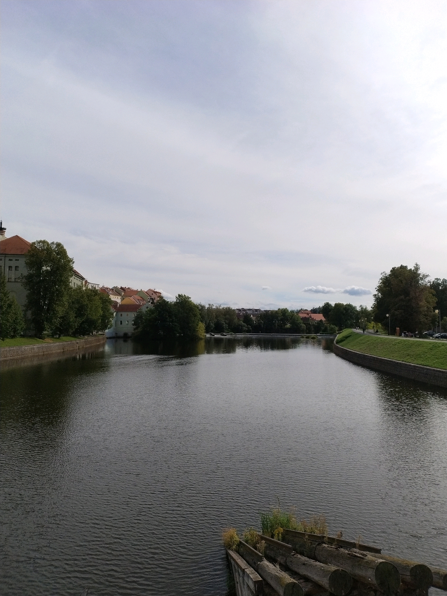 The image depicts a wide river with dark, rippling water, likely the Otava River in Písek, Czechia, given the architectural style of the buildings on the left bank. On the left, a stone embankment with grass borders the river, leading to a large, light-colored building with a red-tiled roof, characteristic of historical European architecture. Adjacent to this are several other buildings with red roofs, interspersed with lush green trees.
The right bank of the river features a manicured, grassy embankment with a stone retaining wall. A paved walkway runs along this embankment, where a few individuals are visible strolling, and parked cars are present. Mature trees, some with full foliage, line this side of the river.
In the foreground, several large, weathered logs are stacked horizontally, forming a rustic barrier or mooring. Some vegetation is growing between the logs. The sky above is overcast with a pale, diffused light, suggesting a cloudy day. The overall scene conveys a tranquil urban riverside landscape with historical elements.