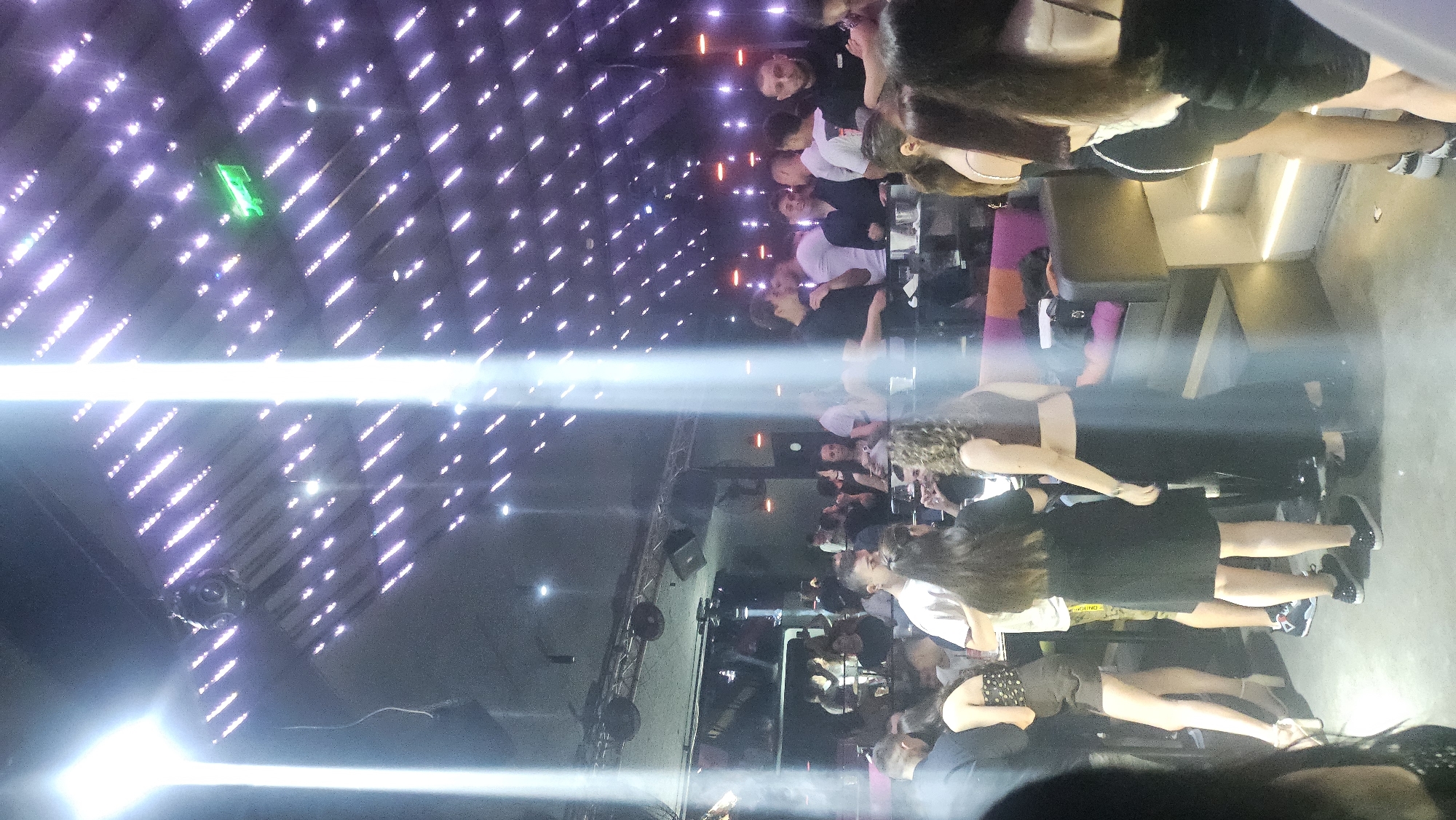 The image displays an interior view of a crowded nightclub or lounge, identified as being in Varna, Bulgaria. Strong, vertical beams of white light emanate from an overhead source, creating prominent lens flares. The ceiling features a grid of diagonally arranged, small, rectangular lights, predominantly purple or pink, and a green illuminated exit sign is visible in the upper right. A round, multi-lensed fixture, likely a disco light or security camera, is suspended from the ceiling in the upper left.
The floor is a light-colored, possibly concrete, surface. Numerous individuals, primarily young adults, are scattered throughout the venue, standing in small groups and engaging in conversation. Clothing includes casual club wear such as dark dresses, crop tops, t-shirts (both white and black), dark trousers, patterned shorts, sneakers, and high heels. Several individuals are holding drinks.
In the mid-ground, small tables hold drinks and glasses. Towards the right, a seating area with multi-colored upholstered benches in shades of purple, pink, and orange is visible, accessible by two illuminated steps. In the background on the left, a dimly lit bar area can be seen with bottles and equipment, where additional patrons are gathered. Overhead, a truss system supports speakers and other event lighting equipment, suggesting a performance area or DJ booth. On the far right, a man wearing a black shirt with a visible white logo, possibly "SESOM," is present.