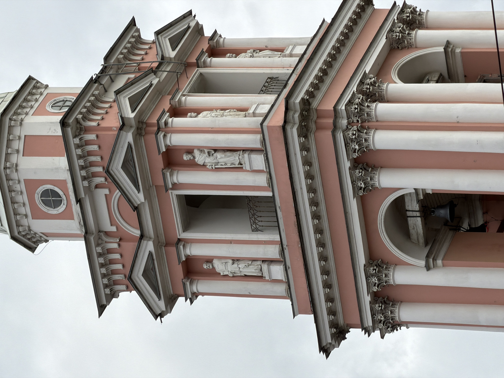 The image shows a close-up, low-angle view of the pink and white bell tower of Saint Nicholas Naval Cathedral in Saint Petersburg, Russia. The tower features classical architectural elements, including multiple levels with arched openings, white Corinthian columns, and statues of figures in togas. A large bell is visible hanging in an archway on the lower level. The sky is overcast, and there is a metal ladder leaning against the side of the tower near the top. The building exhibits ornate decorative molding and cornices, with small circular windows on the upper sections.