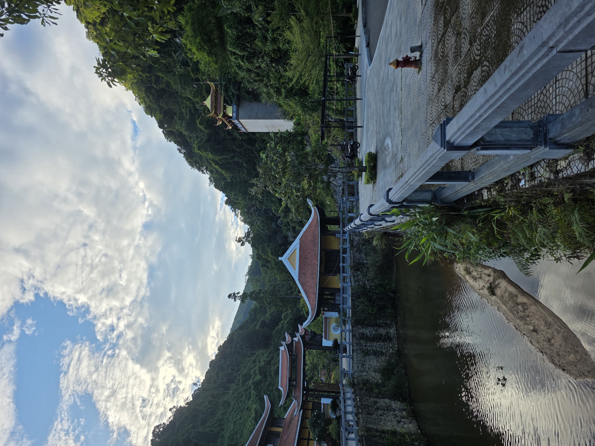 The image depicts a serene temple complex nestled amidst lush green hills. A calm body of water with a small sandbar in the center reflects the cloudy sky and surrounding foliage. Traditional Vietnamese architectural elements are visible, including ornate roofs and pagodas. A stone bridge with a railing separates the water from a paved courtyard. In the distance, a multi-story tower with a gilded roof stands against the hillside. Several motorcycles are parked in the courtyard, and a red fire hydrant is visible near the foreground. The overall scene suggests a tranquil and spiritual atmosphere, likely a Buddhist temple or a historical site. This image was captured in the Bà Nà Hills area of Đà Nẵng, Vietnam.