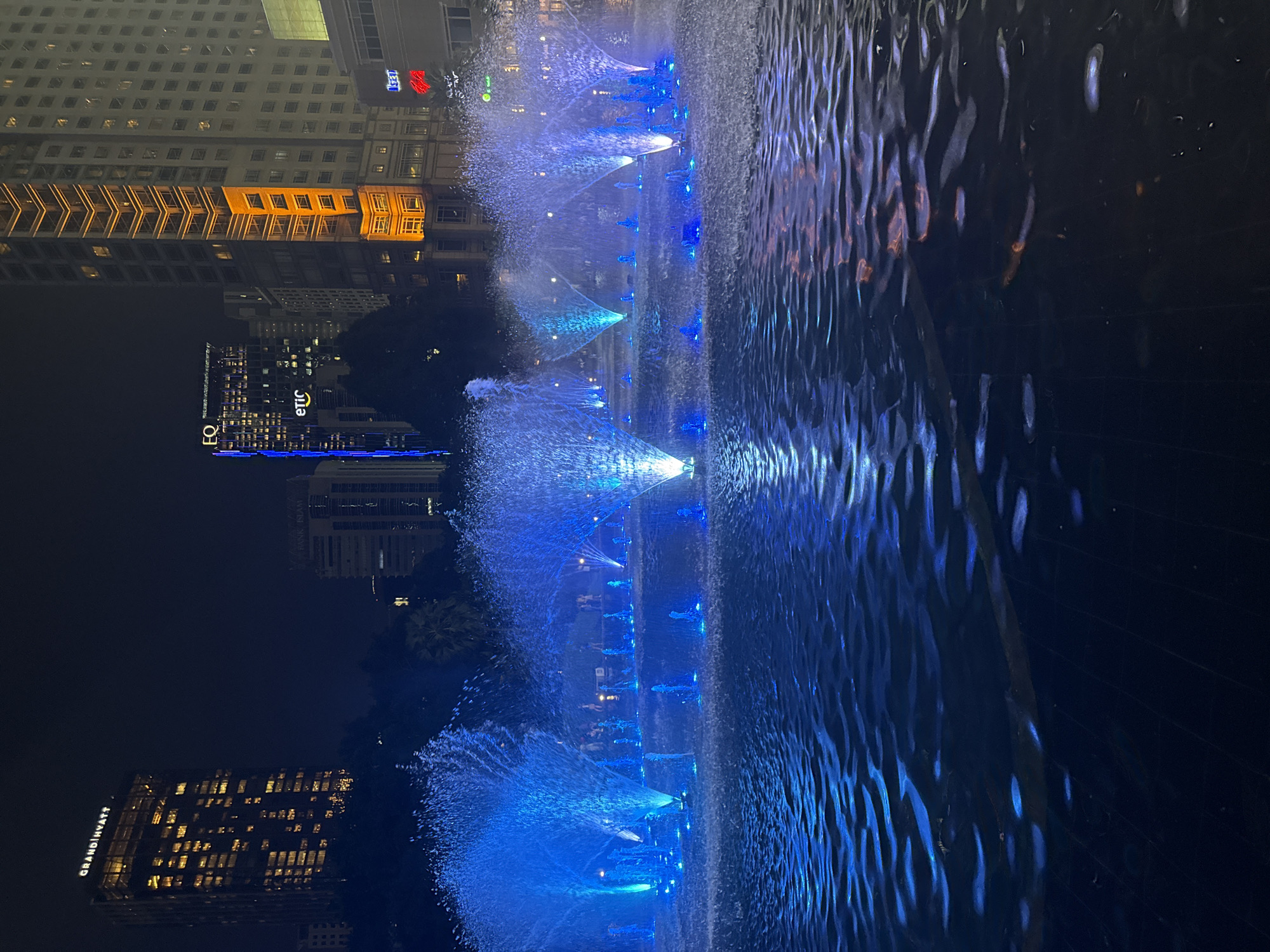 The image shows the Kuala Lumpur City Centre (KLCC) Park Lake Symphony Fountain at night in Kuala Lumpur, Malaysia. The fountain is illuminated with blue lights, and the water jets create a dynamic display. The Petronas Towers and other buildings are visible in the background. The foreground shows the water's surface with wavy patterns, partially reflecting the city lights.