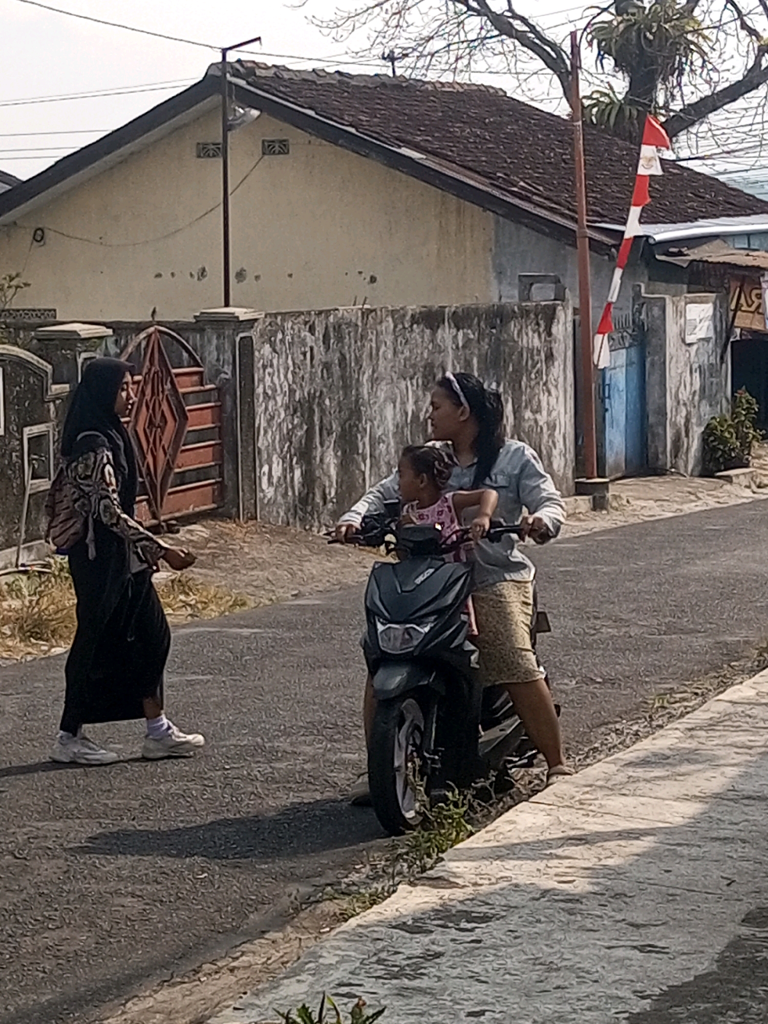 In Krasak, Indonesia, two individuals are present. On the left, a person wearing a black hijab and a patterned top walks along the asphalt road, carrying a backpack. To the right, two individuals, possibly mother and child, are on a dark-colored scooter. The adult, wearing a light-colored jacket and a patterned skirt, is operating the scooter, while a child in a pink shirt is seated in front of them. The scooter is positioned on the road, facing right. In the background, a building with a tiled roof and a weathered wall is visible. A flagpole with a red and white Indonesian flag is also present. The ground is a mix of asphalt and a concrete sidewalk. The overall setting appears to be a residential street.