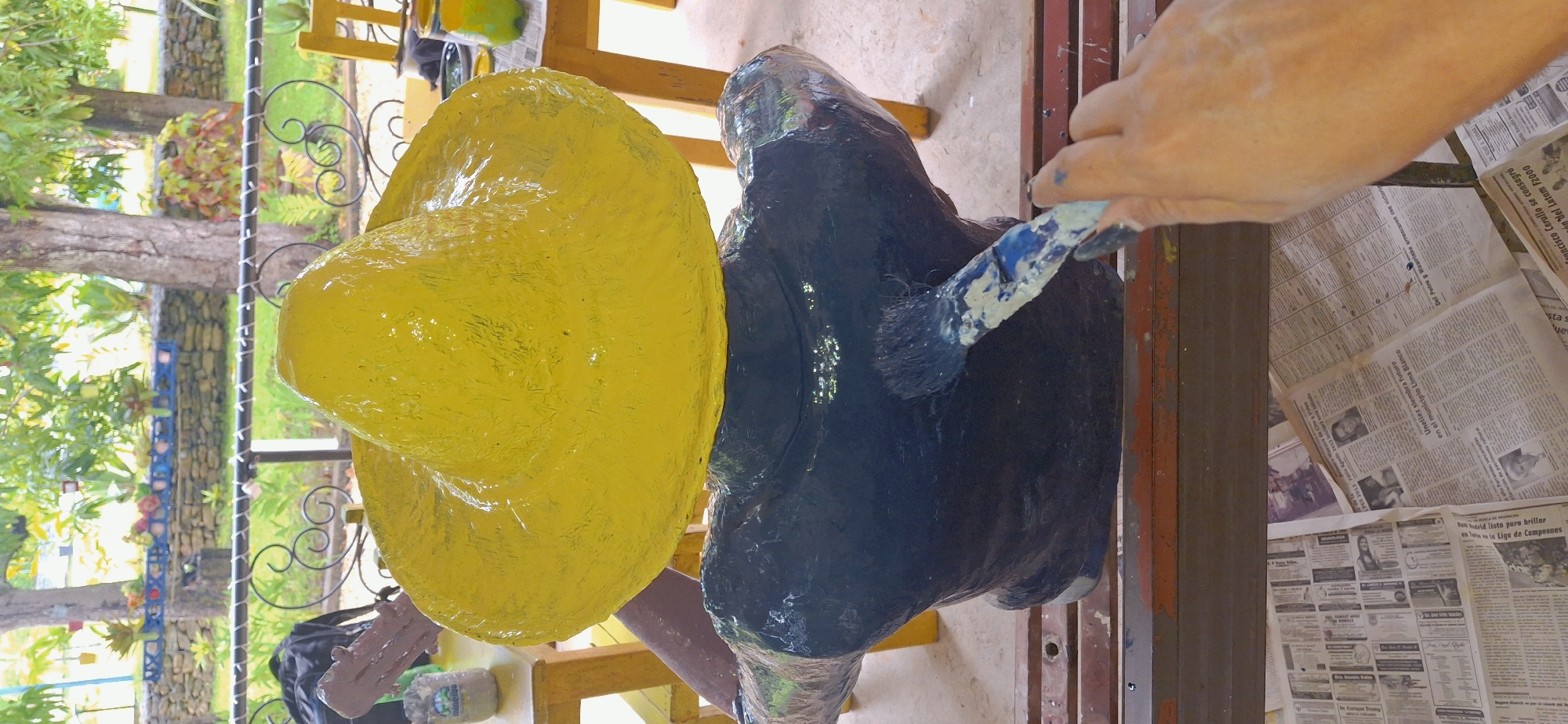 A person's hand is visible painting a dark blue shirt on a statue. The statue has a large, yellow sombrero on its head, which appears to be made of a textured material. The painting is taking place outdoors, with trees and foliage visible in the background. In the distance, a railing separates the area from what appears to be a garden with potted plants and stone walls. A yellow chair and a table are also visible, suggesting a dining or relaxation area. The ground is covered with newspapers, which are likely being used as a protective covering. The overall impression is of an artistic endeavor taking place in a semi-outdoor environment.