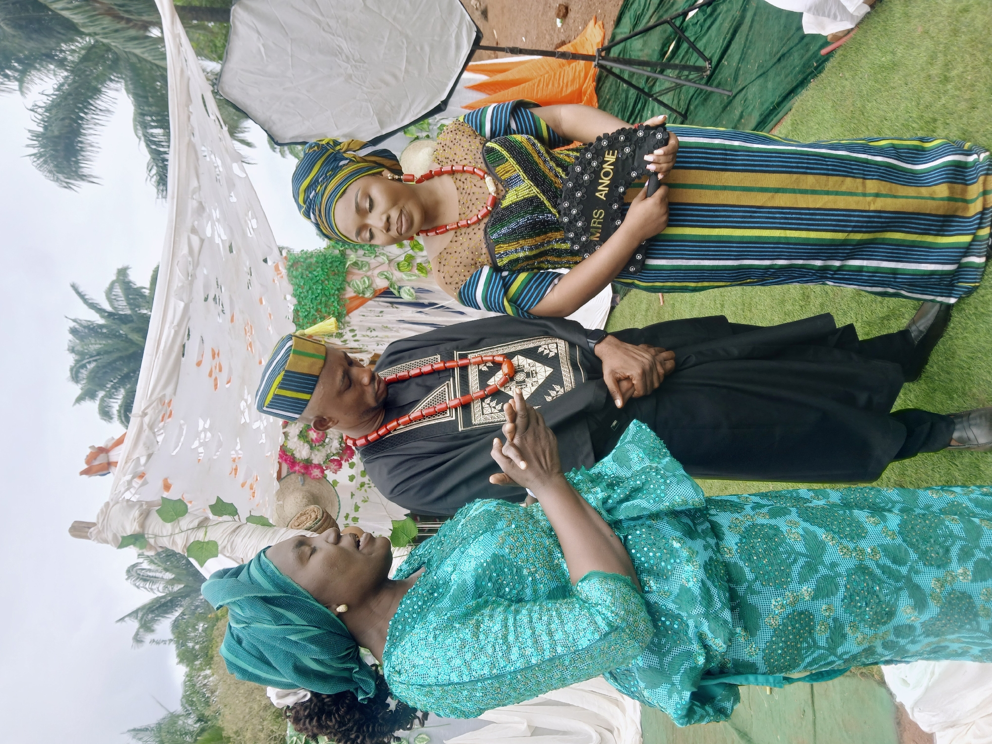 Three individuals are present in an outdoor setting. A woman stands wearing a striped dress and headwrap, a red necklace, and holding a fan that reads "MRS ANONE." A man sits dressed in black with a decorative pattern, a matching hat, and a red necklace. Another woman in a green, sequined dress and headwrap stands next to him. The environment appears to be a yard with a decorated backdrop suggesting a wedding or celebratory event in Ogbonicha, Nigeria. A photography light stand is visible.