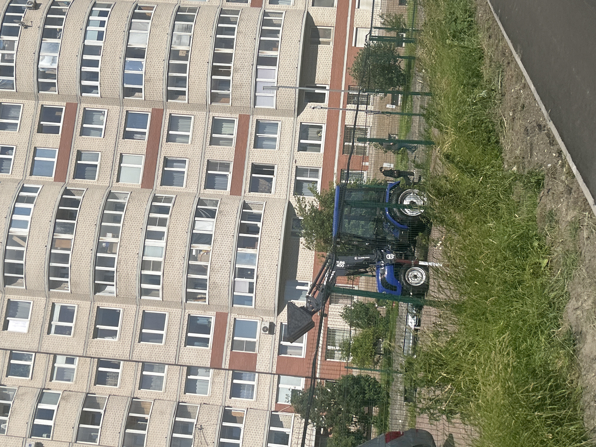 A blue tractor with a raised bucket is parked behind a fence in front of a light-colored brick apartment building in Saint Petersburg, Russia. The building has many windows and balconies, and a red brick accent line runs horizontally across its facade. Tall green grass grows in front of the fence, obscuring the lower part of the tractor. Two individuals, one in dark clothing and another in lighter clothing, are visible standing near the tractor behind the fence. A gray vehicle is partially visible on the left side of the image. The ground in front of the grass appears to be dirt and gravel.