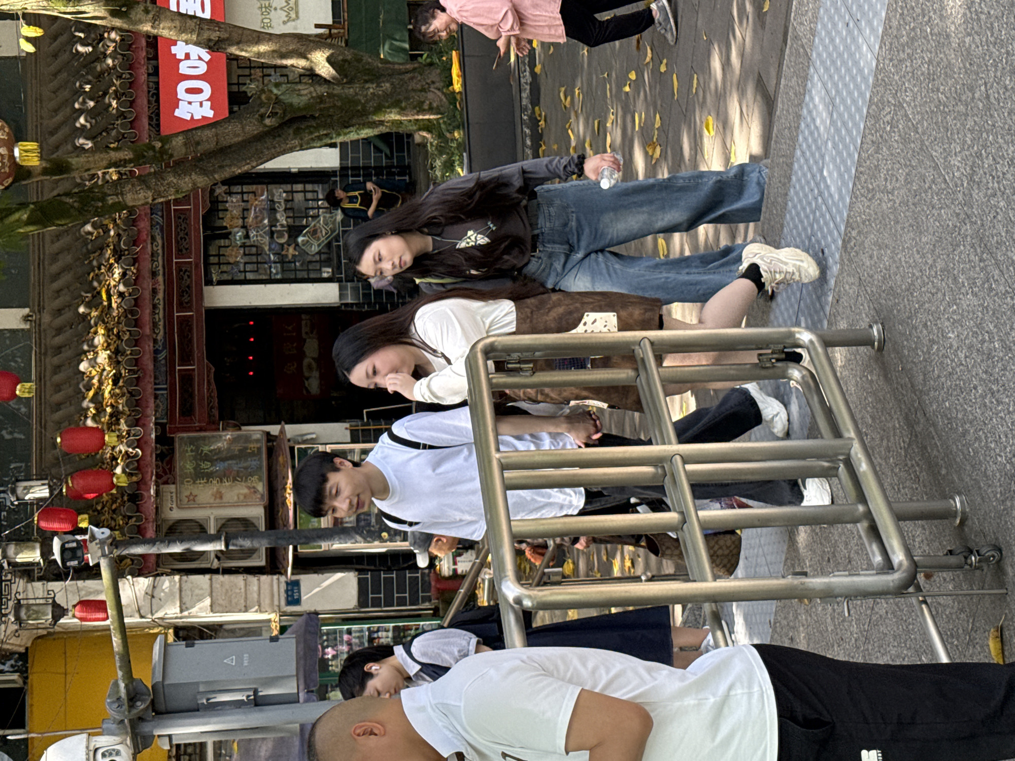 Here is a forensic description of the image:
The image depicts a street scene in Jiulongpo District, China. Several people are visible in the frame, including three women and two men. The women are wearing casual clothing: one has a black hoodie and jeans, another has a white shirt and a brown skirt, and the other has a white shirt with a navy dress. One of the men wears a white shirt and black sweatpants. The other man wears a white shirt and jeans. The individuals appear to be walking or standing near a silver metal barrier.
The environment includes a building with traditional Chinese architectural features, red lanterns, and signs with Chinese characters. There are air conditioning units mounted on the exterior of the building. A tree is present with leaves on the ground. The ground is made of light grey concrete and has a tactile paving section. A traffic signal pole is visible on the left side.
