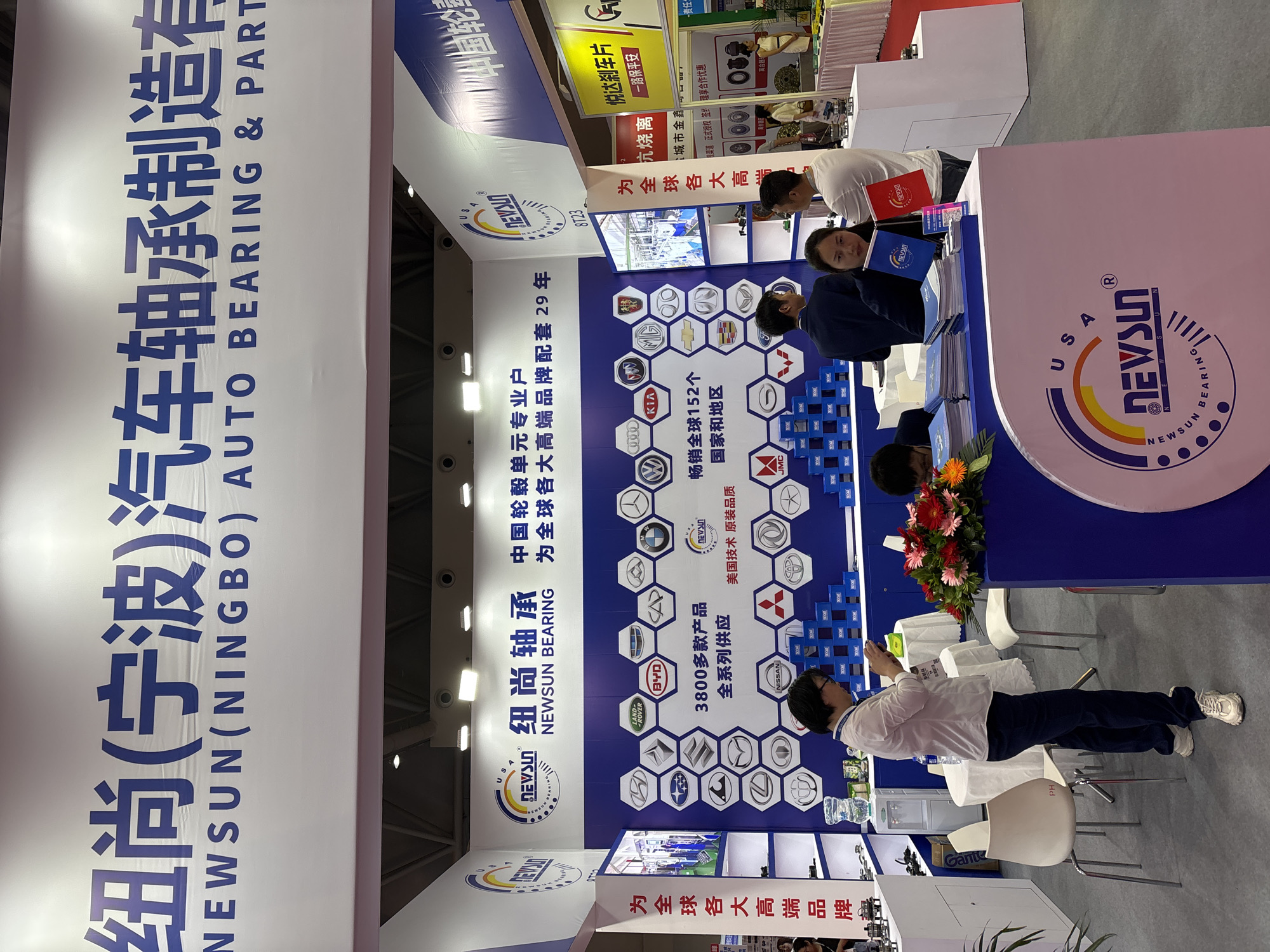 A trade show booth displaying "NEWSUN (NINGBO) AUTO BEARING & PART," located in the Liangjiang New Area of Chongqing, China, showcases bearings and auto parts to attendees. The booth has banners with the company name in Chinese and English. The trade show booth has a table with people looking at the parts. There are many logos of car brands on the back wall, with the company selling to many countries. A person is taking a picture of the stand.