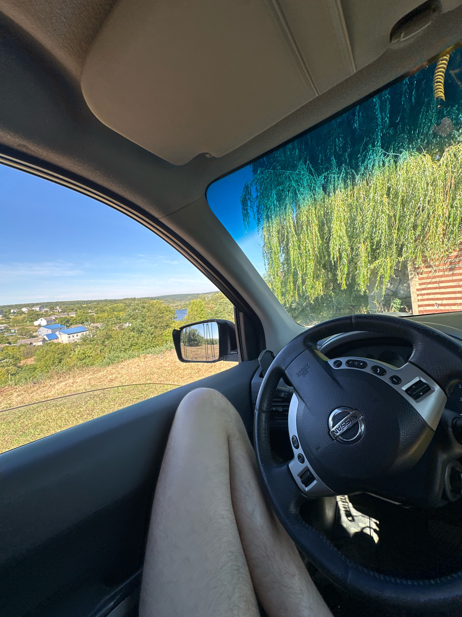 This is a first-person perspective image taken from inside a car. The shot is framed by the car's interior, showing the steering wheel with the Nissan logo and various buttons. To the left, a person's legs are visible, resting against the car door. Through the open car window, a picturesque rural landscape unfolds, featuring green rolling hills dotted with houses, a body of water, and lush trees. The sky is a clear blue. To the right, a wall made of red brick is visible, partially obscured by hanging green foliage. The image captures a moment of travel and scenic enjoyment in what appears to be a rural setting, possibly on the outskirts of Makiv, Ukraine, given the village-like appearance and rural landscape.