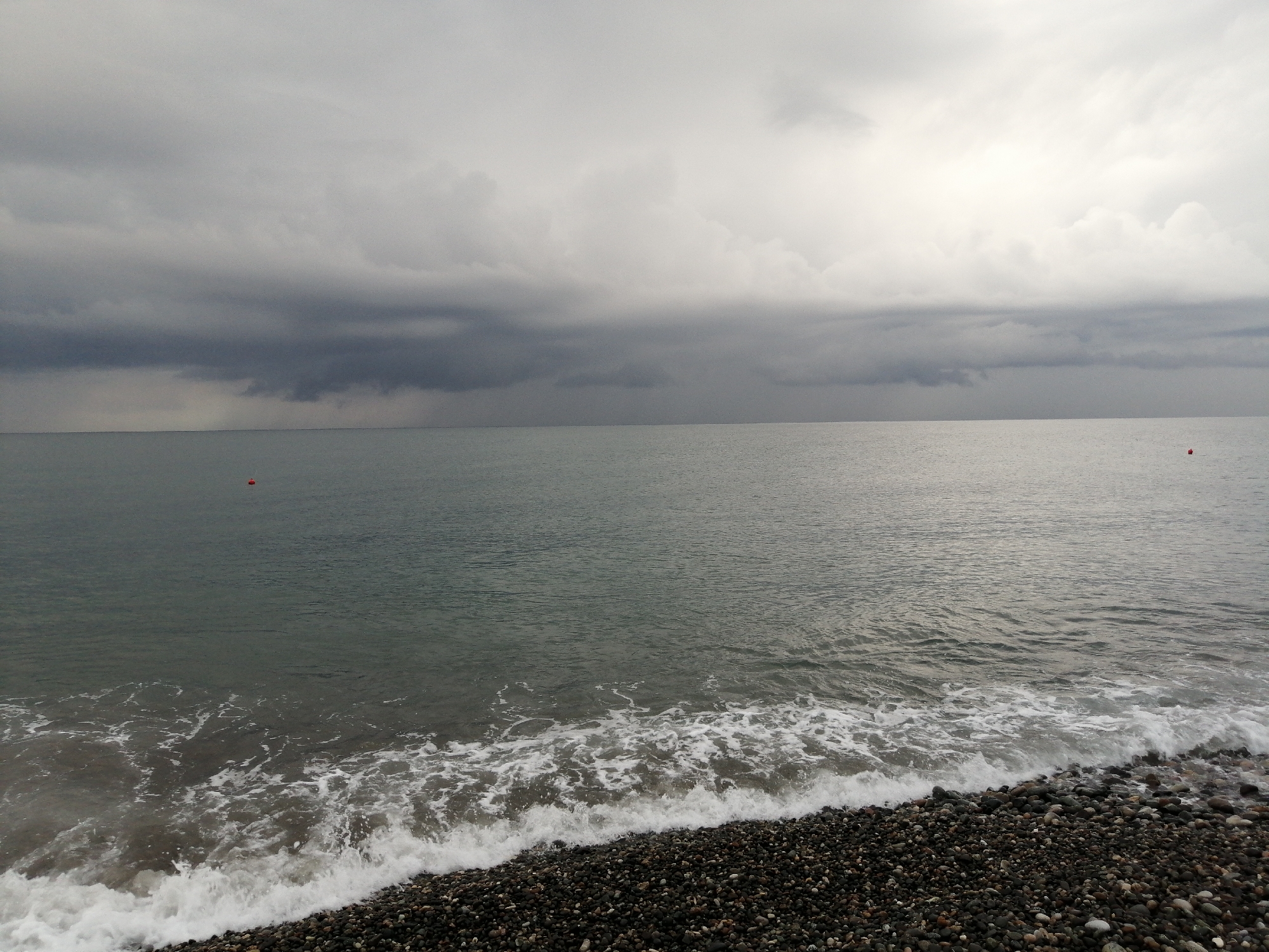 A wide shot shows a pebble beach in Batumi, Georgia, meeting a calm sea under an overcast sky. The sea is a muted green, with gentle waves lapping at the shore. The beach is covered in dark, rounded pebbles. In the distance, two red buoys are visible on the water. The sky is dominated by heavy, dark clouds, with a brighter patch of sky to the right, suggesting the sun may be attempting to break through. The overall atmosphere is subdued and slightly melancholic.