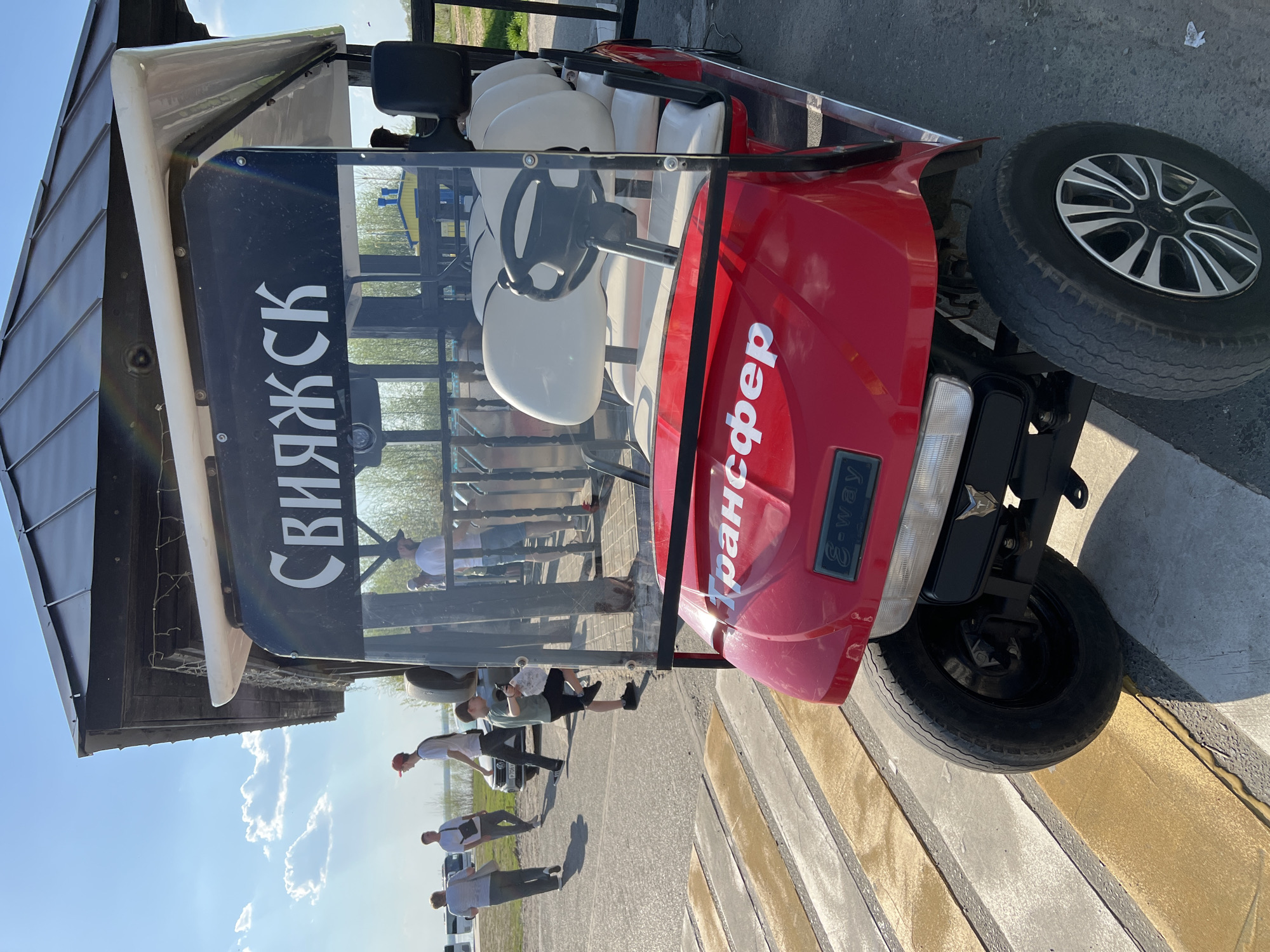 The image depicts a red golf cart with white seats parked on a gray asphalt road. The cart has the word "Трансфер" in white Cyrillic letters on its front hood and "Свияжск" (Sviyazhsk) in white Cyrillic letters displayed vertically on the front windshield. A small "E-way" emblem is visible on the cart's front right side. In the background, there is a building with a dark roof and a partially obscured wooden structure. Several people are standing or walking in the background near the building. The sky is blue with scattered white clouds. The ground is lined with alternating gray and yellow stripes, resembling a crosswalk. The photograph appears to be taken during daylight hours in Sviyazhsk, Russia.