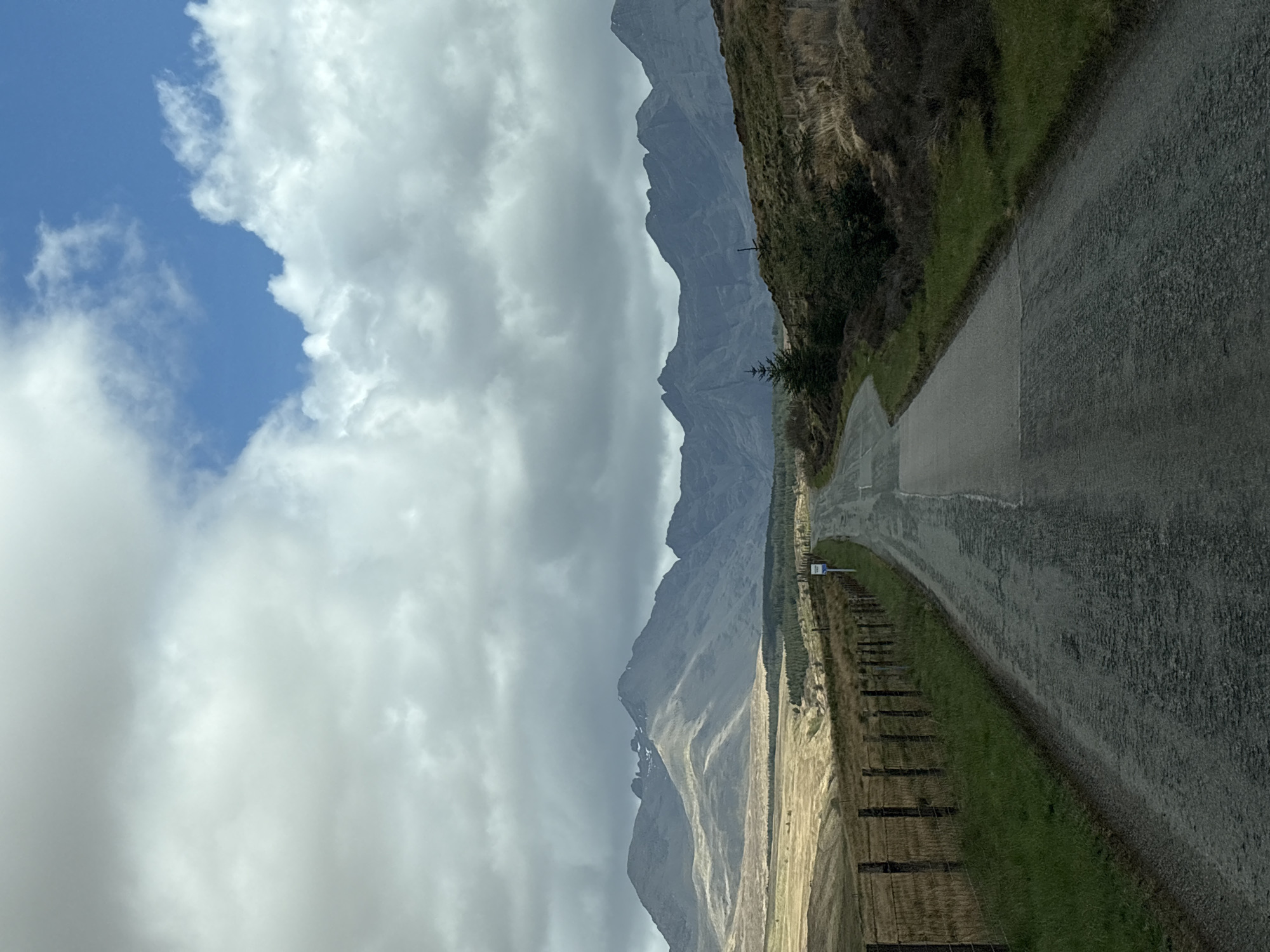 A road in the Isle of Skye, United Kingdom, is depicted under a cloudy sky. The road runs vertically along the right side of the image. To the left of the road, there's a grassy area, then a field, and beyond that, a mountain range. The sky is predominantly filled with grey clouds, with a small patch of blue sky visible in the upper left corner.