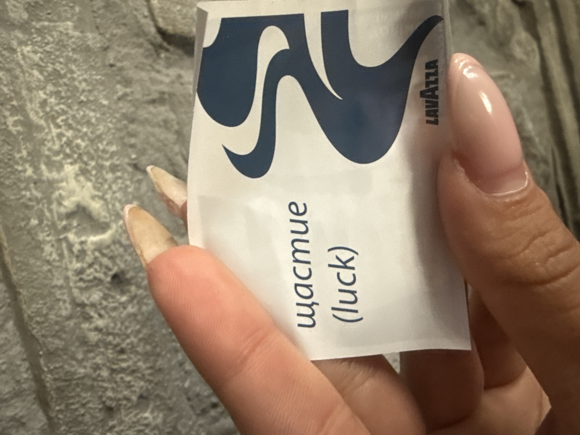 A hand holds a small, white packet of Lavazza sugar. The packet has a blue Lavazza logo and the Russian word "счастье" (luck) printed on it. The background is a grey, textured surface. This image was taken in the city of Pleven, Bulgaria.