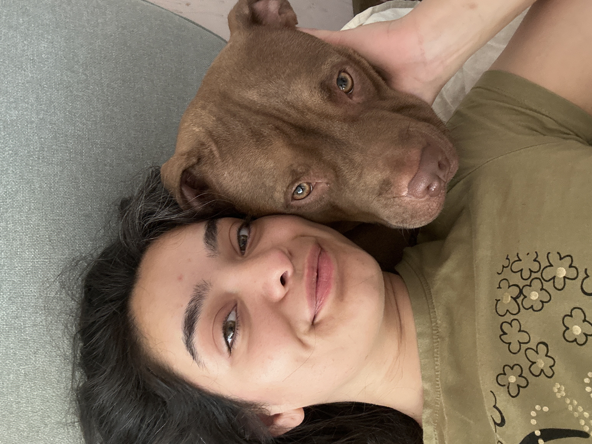 The image shows a close-up selfie of a young woman and a brown pit bull dog lying together. The woman has dark hair and is smiling at the camera. The dog's head is resting on her shoulder, and she is gently stroking its head. The background appears to be a couch with a textured gray fabric. The scene is one of affection and companionship between a human and her pet. There is no information available that indicates the location is in the city of Malevo, Bulgaria.