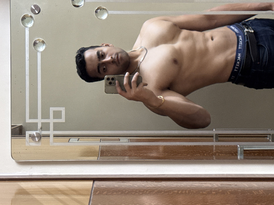 A shirtless man, angled in the frame, takes a selfie in a mirror. He's wearing jeans and a dark belt with a visible logo on the waistband. The man has dark hair and a gold chain necklace. He is holding a smartphone with his left hand. The mirror is decorated with a geometric pattern and crystal-like embellishments. The background appears to be a neutral wall and a wooden surface.