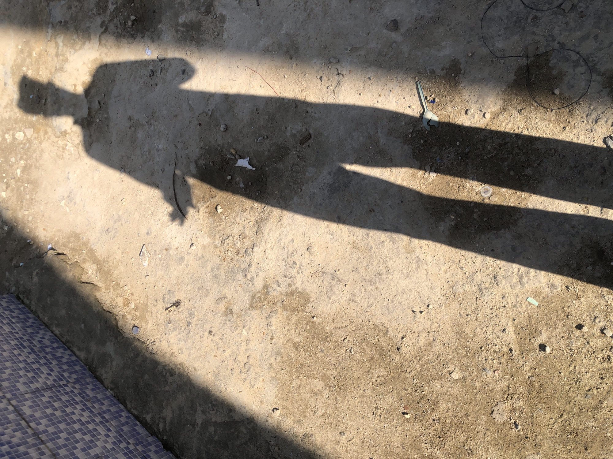 The image shows the shadow of a person cast on a rough, sandy ground. The shadow is long and stretches diagonally across the frame, indicating the presence of a strong light source, likely the sun. The person's shadow appears to be wearing a shirt and pants. In the background, there is a section of a wall covered in blue and white checkered tiles. The ground is littered with small stones, debris, and some scattered pieces of paper. There is also a pale blue tool with a curved handle lying on the ground to the right of the shadow. The overall scene suggests an outdoor urban environment, possibly under construction or in a state of disrepair, located in the city of Ado, Nigeria. The lighting and the shadow suggest it is daytime.