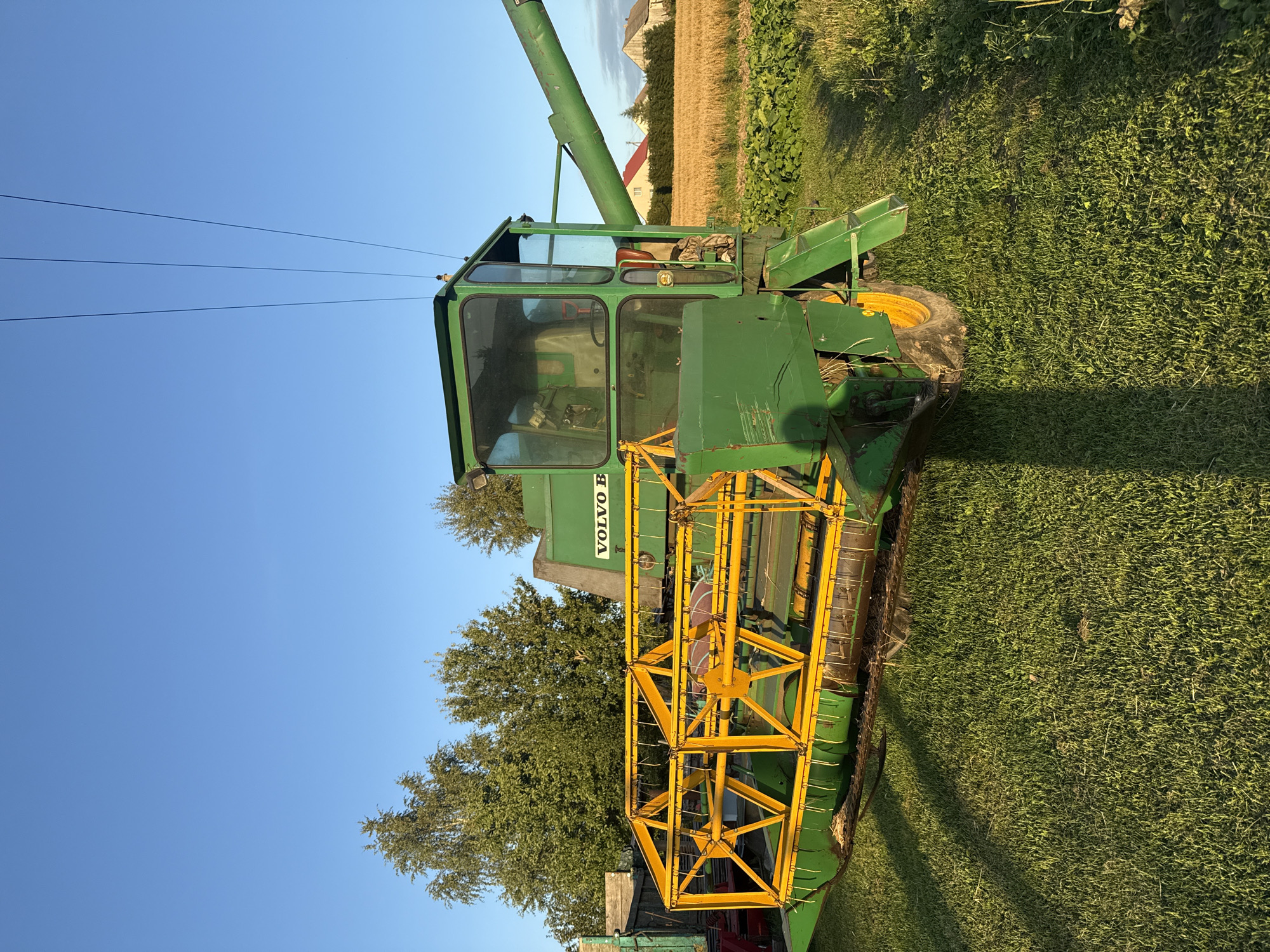 In Vikraičiai, Lithuania, a green Volvo B combine harvester sits parked on a grassy field under a clear blue sky. The combine, adorned with a yellow header, faces left, its main body visible from the side. Behind it, a field of golden crops stretches towards a few houses in the distance. Trees line the left edge of the frame, with a wooden structure partially visible. The sun casts long shadows across the grass, indicating it is late afternoon or early evening.