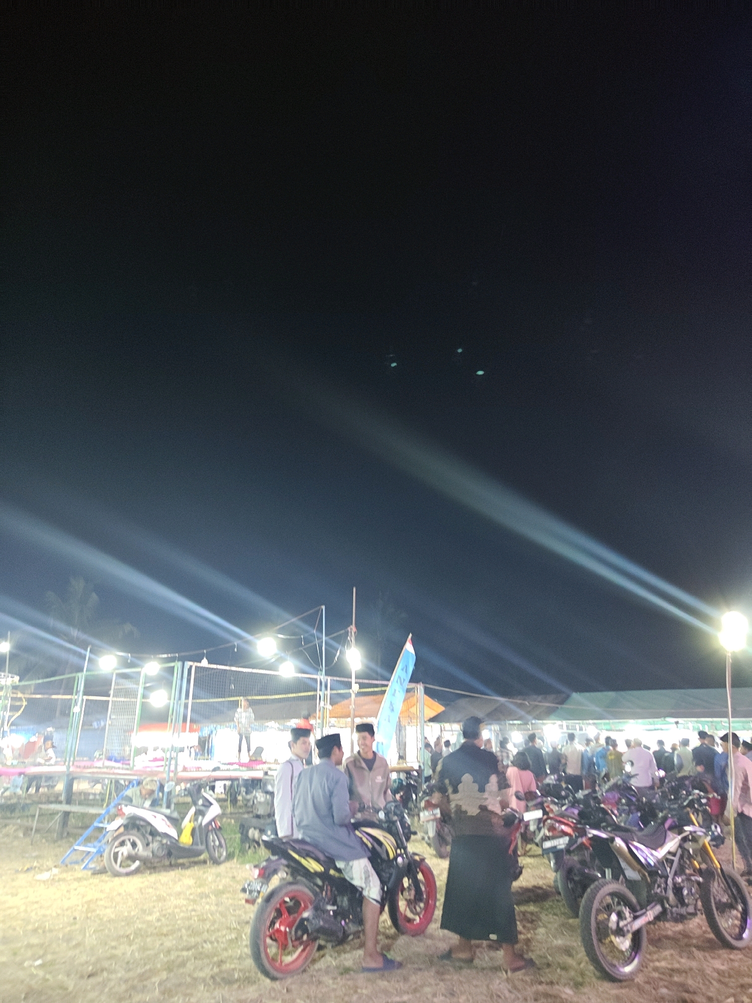 The image depicts a nighttime outdoor event in Suling Wetan, Indonesia. Several men are gathered, some are standing and talking, while others are seated on motorcycles. In the foreground, two men are on motorcycles. One man, wearing a gray shirt and white shorts, is seated on a black motorcycle with yellow accents and red rims. The other man, wearing a black and tan patterned jacket and a black skirt-like garment, is standing beside a black motorcycle. In the background, a crowd of people is visible, as are numerous motorcycles parked. Several bright lights illuminate the scene, casting long beams across the dark sky. There are also structures that appear to be temporary stalls or stages in the background, suggesting a market or festival setting. The ground is covered with dry grass and straw.