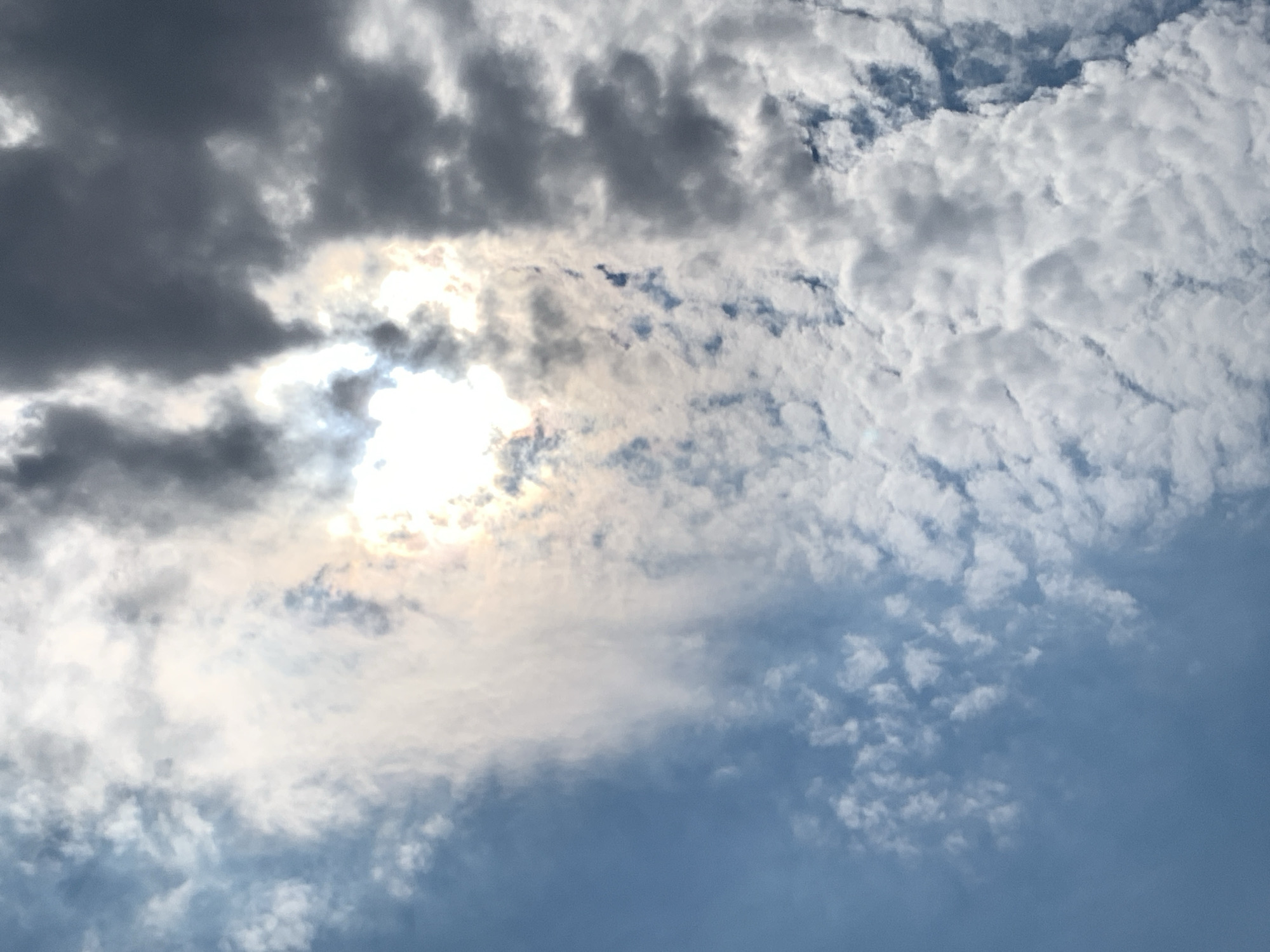 The image displays a sky filled with clouds. The sun is visible, peeking through a gap in the clouds, causing a bright, luminous effect in the center of the frame. The clouds are varied in texture and color, ranging from bright white to dark gray, suggesting a mix of conditions, possibly indicating an approaching storm or a transition in weather. The blue sky is visible in the lower left quadrant, interspersed with patches of thinner, wispy clouds. There are no people, subjects, or objects visible in the image, nor are there any actions or interactions depicted. The image appears to be a landscape shot focused solely on the atmospheric conditions of the sky. It is not possible to determine the specific location within Khazar Raion, Azerbaijan, from this image alone, as it only captures the sky.