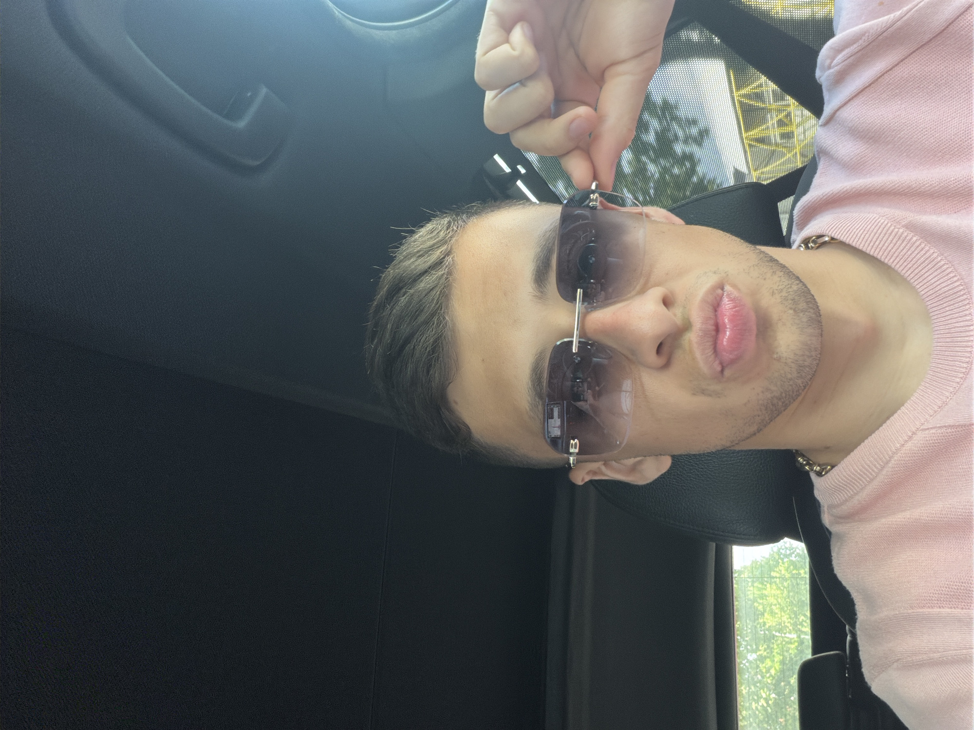 A man is sitting inside a car. He has dark hair and is wearing sunglasses. He is also wearing a pink sweater and a gold chain. The background shows trees and a building. The location appears to be in a city.