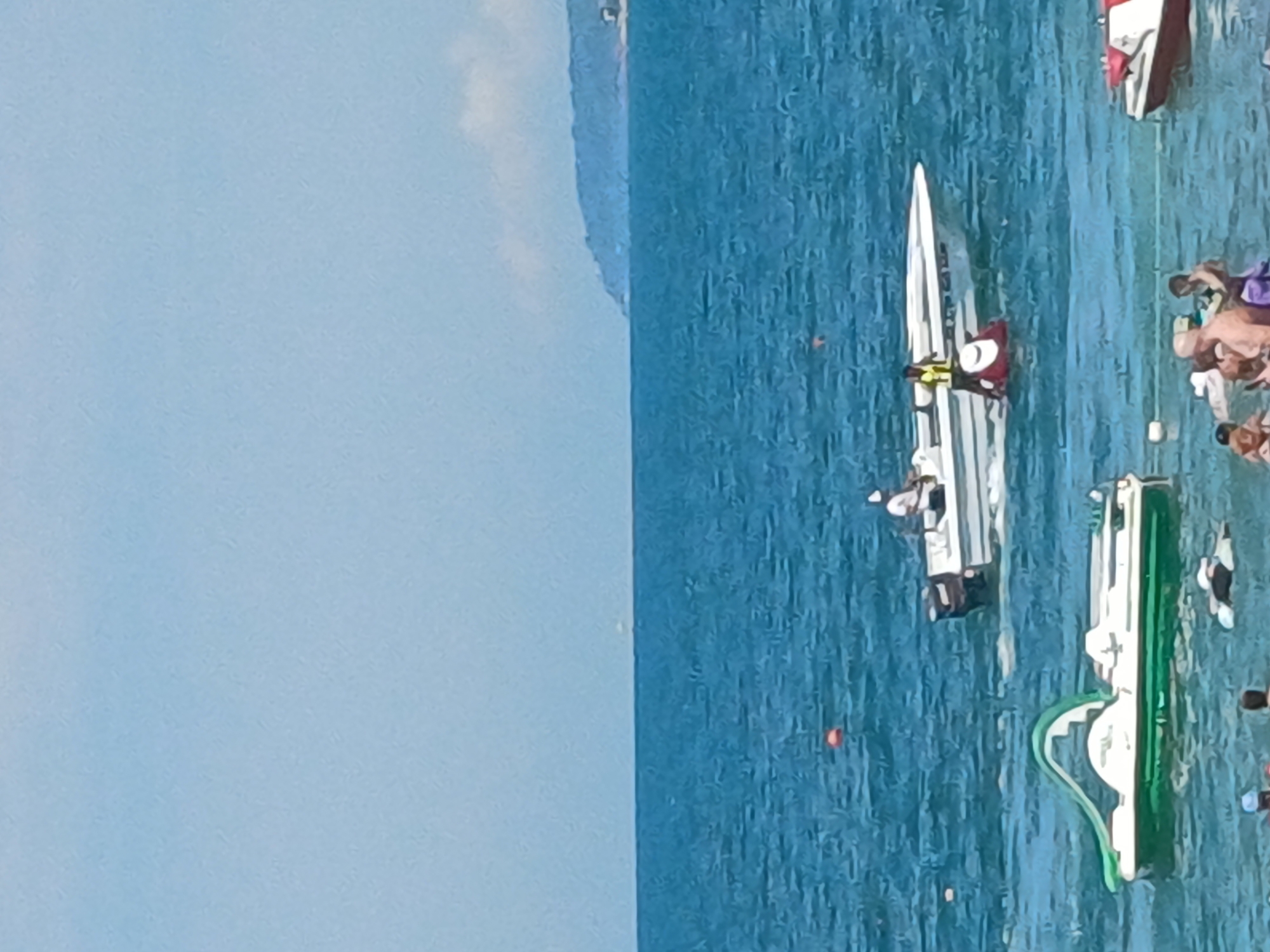 The image depicts a sunny day at a beach in Sochi, Russia, with the Black Sea as the backdrop. Several boats are visible on the water, including a white speedboat with two people onboard and a jet ski rider. In the foreground, a group of people are enjoying the water, with some partially submerged. A green and white pedal boat with a slide is also present, along with other smaller watercraft. The distant shoreline features a tree-covered hill, suggesting the natural beauty of the region. The sky is clear and bright blue, with the sun casting a warm glow on the scene.