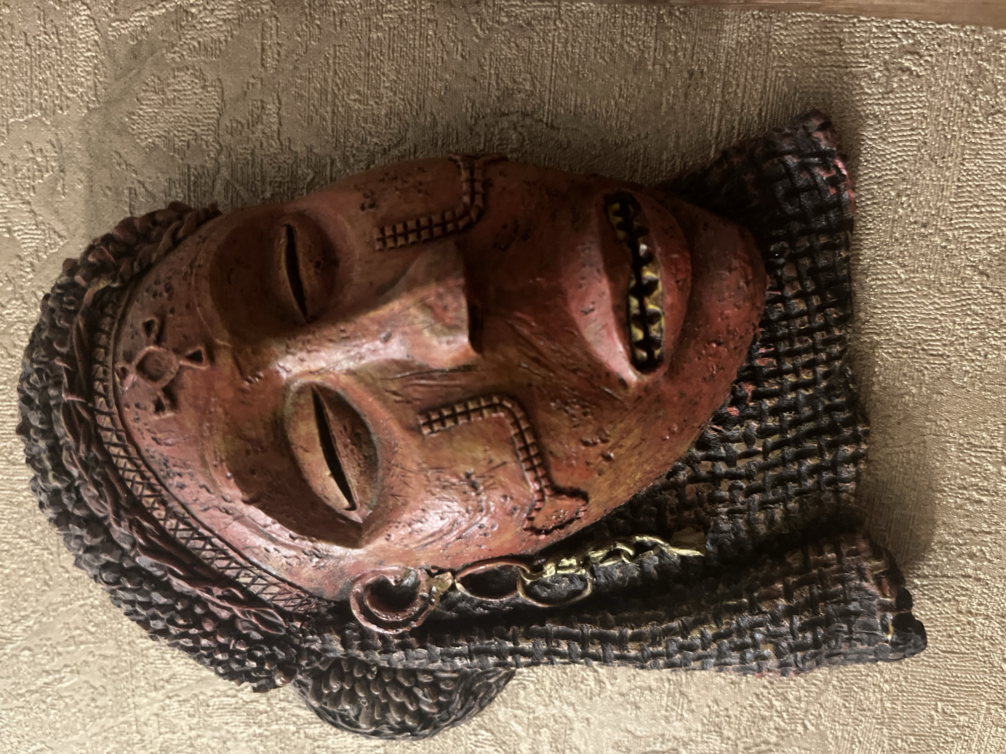 A close-up, slightly high-angle shot shows a textured, reddish-brown tribal mask mounted on a textured, beige wall. The mask depicts a face with closed eyes, an open mouth with visible teeth, and an ornate headdress. A dark, braided hair-like structure frames the face, and a gold-colored earring hangs from the right ear. The mask appears to be made of a hard material, possibly wood or plaster, and exhibits a weathered, aged appearance with visible cracks and indentations. The background wall has a repeating, raised pattern that creates a subtle texture. There are no people or animals present in the image. The location in Vladimirovka, Russia, is not discernible from the visual information provided.