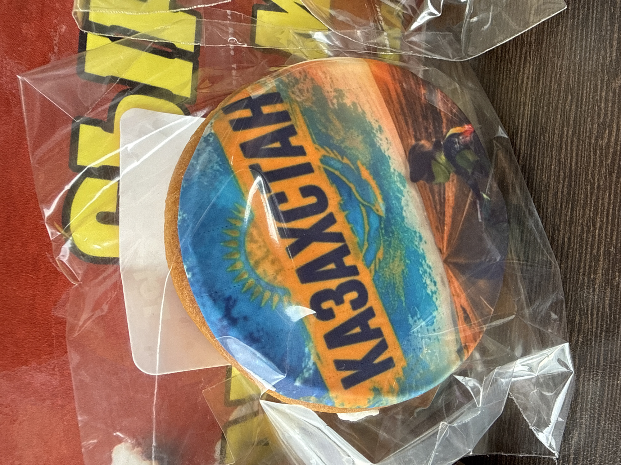 The image shows a close-up view of a round cookie decorated with the flag of Kazakhstan. The cookie is enclosed in clear plastic packaging. In the background, partially visible, are bright yellow letters that appear to be from a sign or packaging, possibly with a red background. The cookie itself features a blue sky with a yellow sun and a silhouette of a person against an orange and brown landscape. The text "ҚАЗАҚСТАН" (Kazakhstan in Kazakh) is prominently displayed in dark blue lettering across the cookie's surface. The cookie is placed on a dark wooden surface. There is no discernible relation to the city of Inkerman, Ukraine.