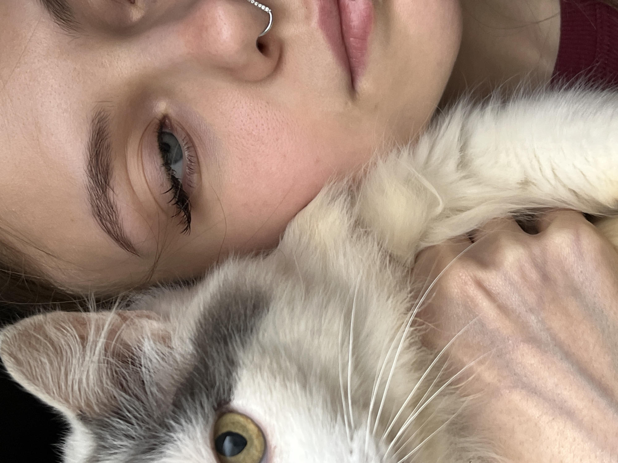 A close-up shot shows a young woman resting her face against a cat. The woman has light skin, defined eyebrows, and a nose ring. Her eye is open and appears to be blue. The cat is white and gray with yellow eyes. The woman's hand is also visible, holding the cat. The setting appears to be indoors. There is no indication of the location being Фоки, Russia.