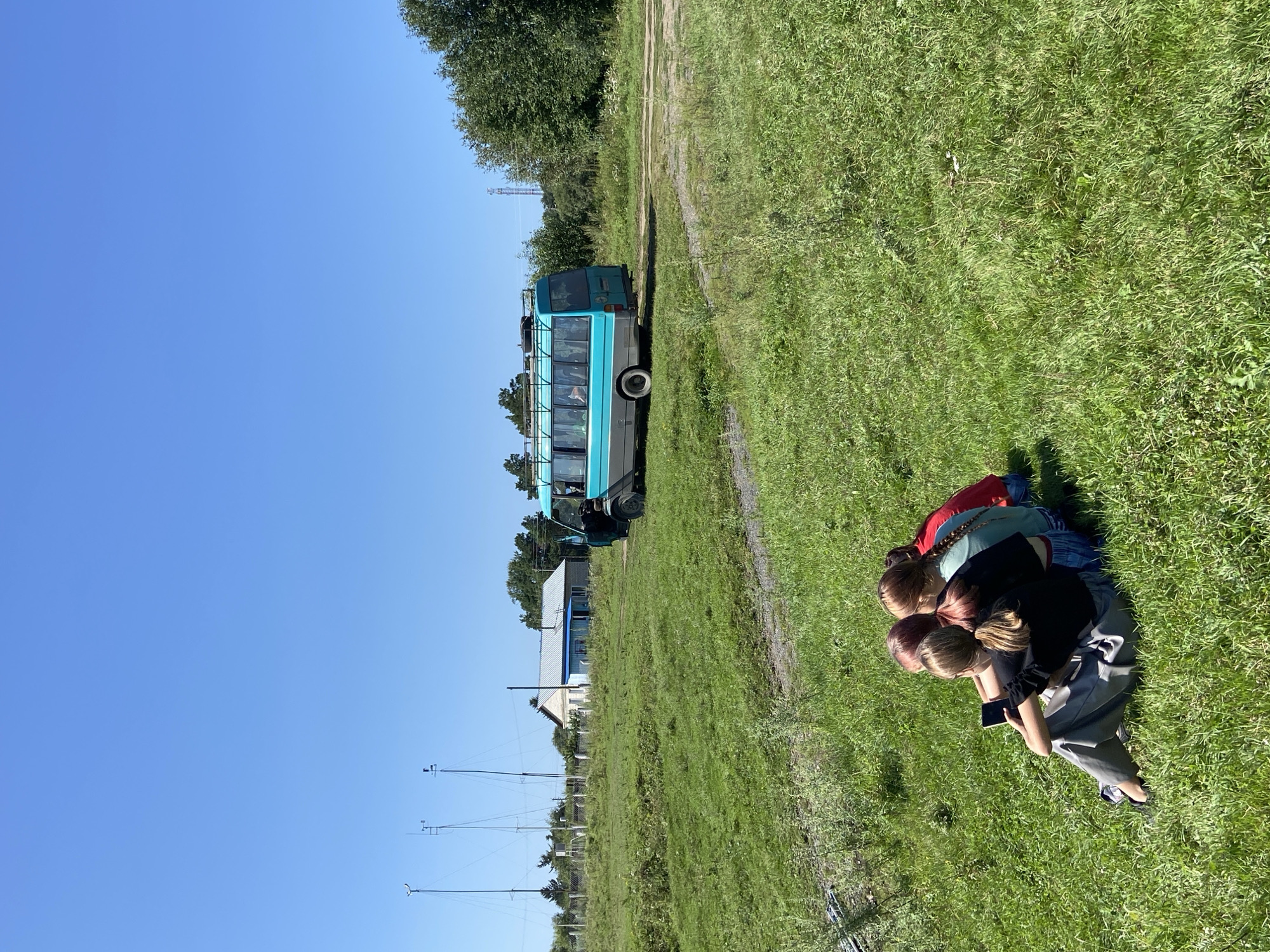 Four young individuals are seated in a grassy field facing away from the camera, with one of them holding a mobile phone. Behind them, a turquoise and gray bus is parked on a dirt path. In the background, a light-colored house with a blue roof and several tall, slender towers are visible, along with trees and a clear blue sky. The environment appears to be a rural or semi-rural area.